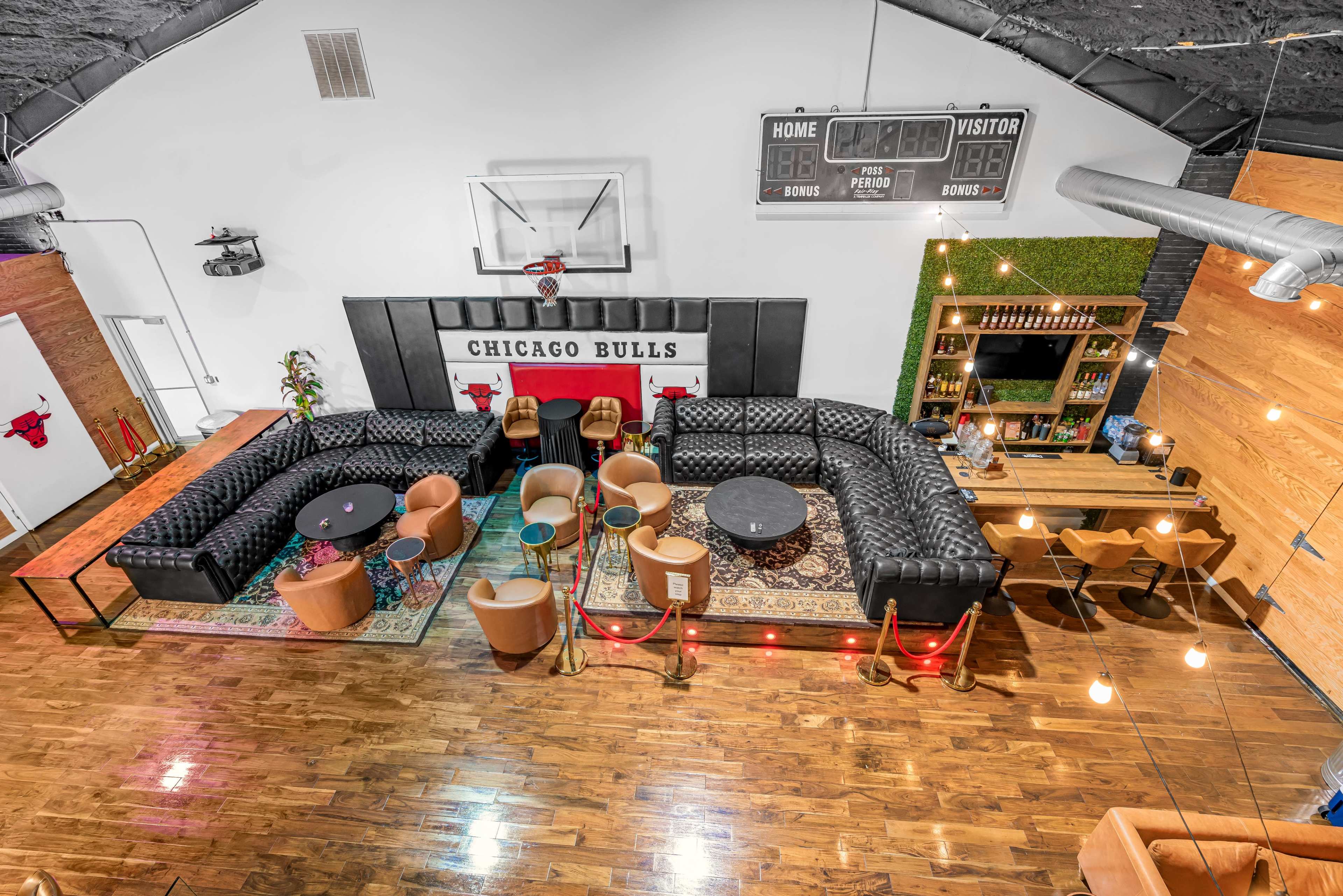 A spacious lounge area featuring a large black leather sectional sofa, wooden flooring, and a bar setup, with Chicago Bulls decor on the walls.