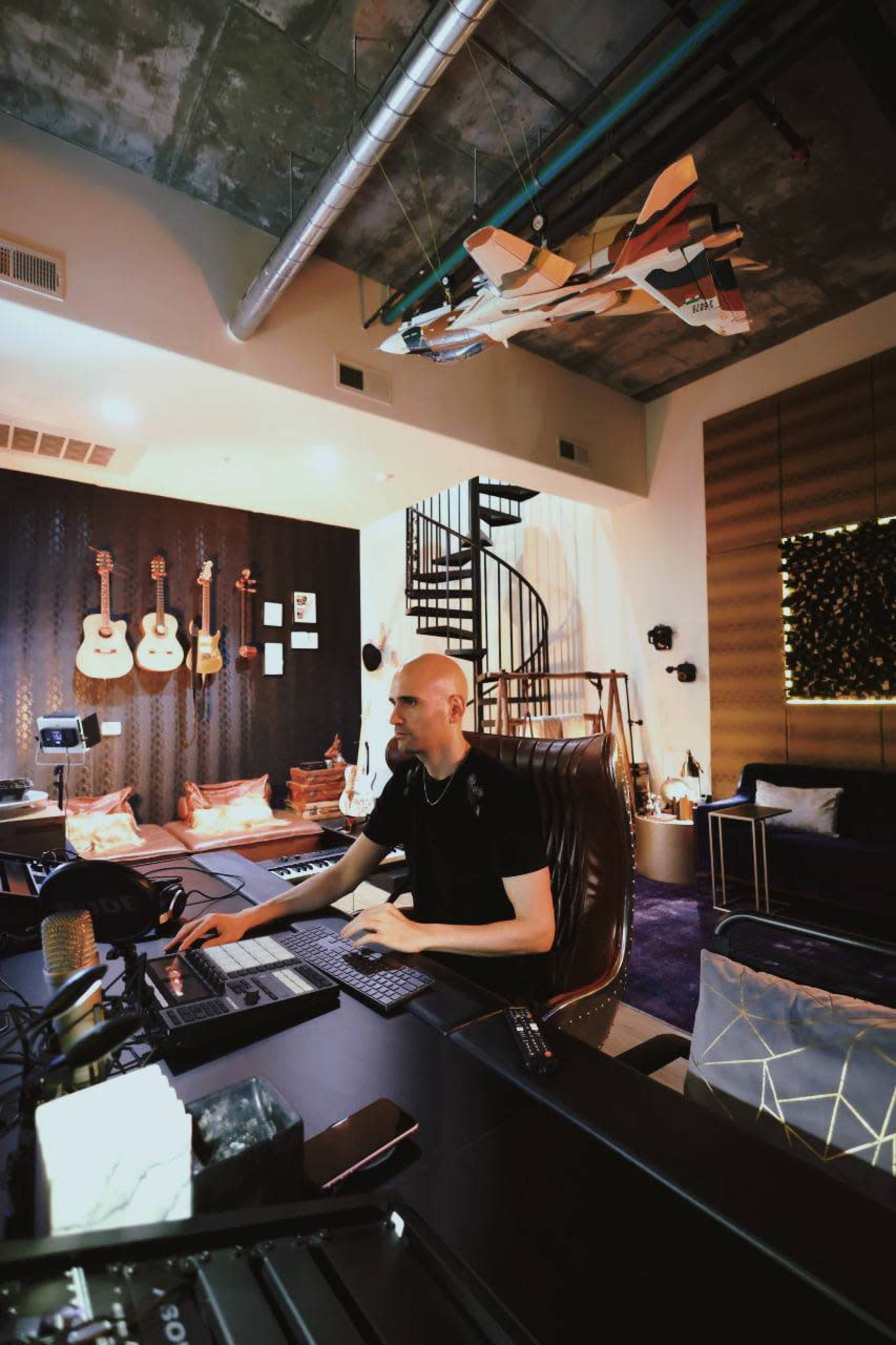 A man sits at a desk with musical equipment in a modern studio featuring guitars on the wall and a spiral staircase in the background.