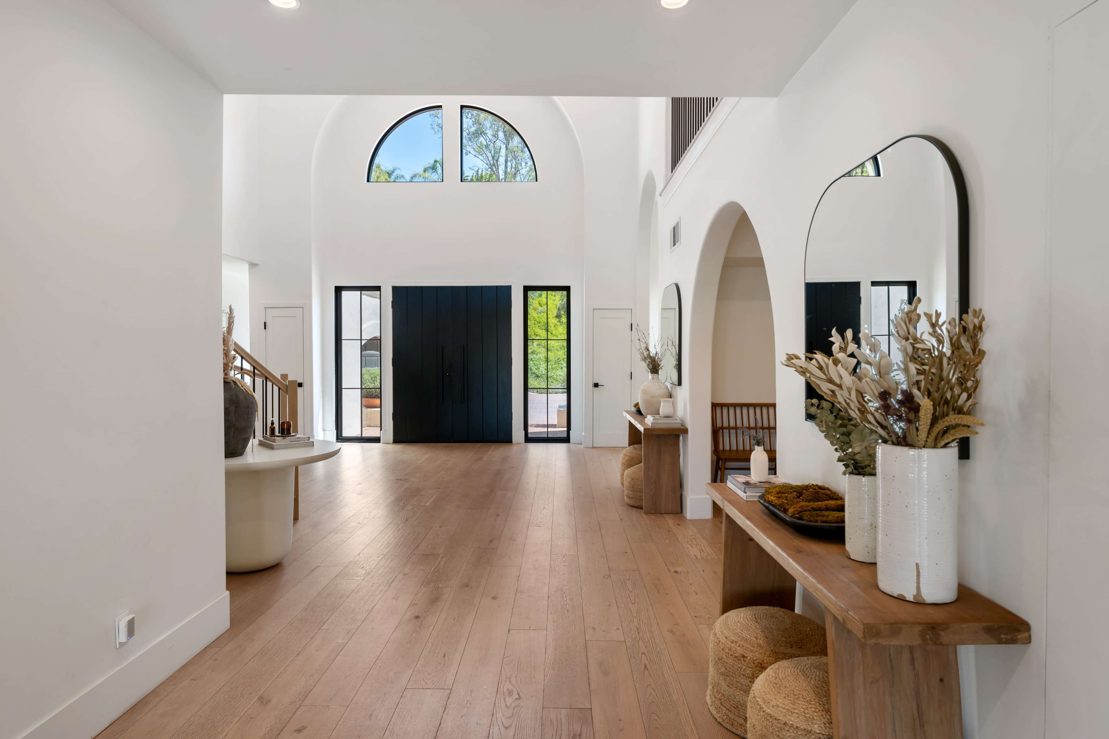 A modern, bright entryway featuring large arched windows, a wooden floor, and a minimalist decor arrangement with a table, vases, and mirrors.