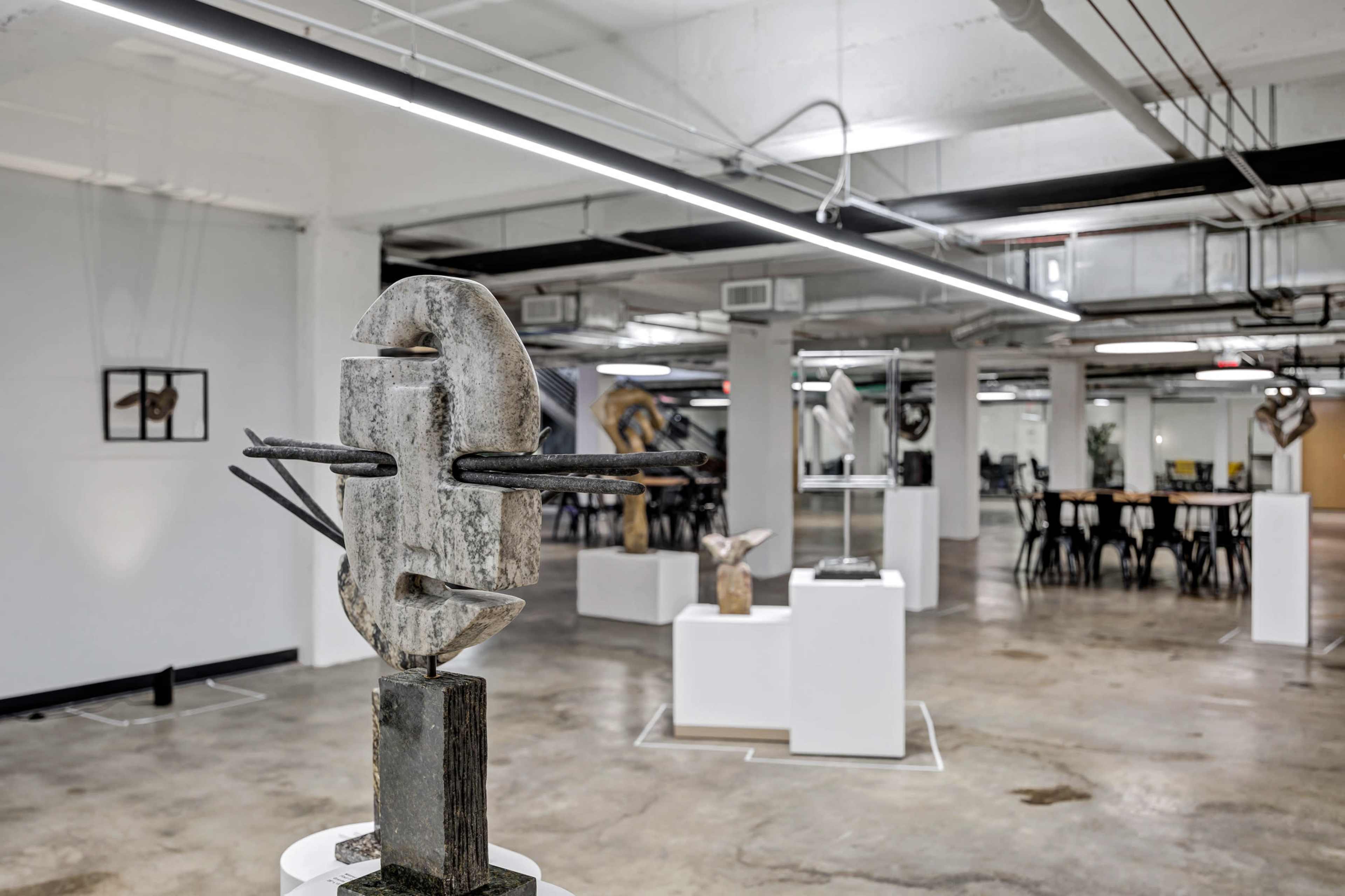 The image shows a modern art gallery with various sculptures displayed on white pedestals and an open floor plan featuring concrete flooring and overhead lighting.