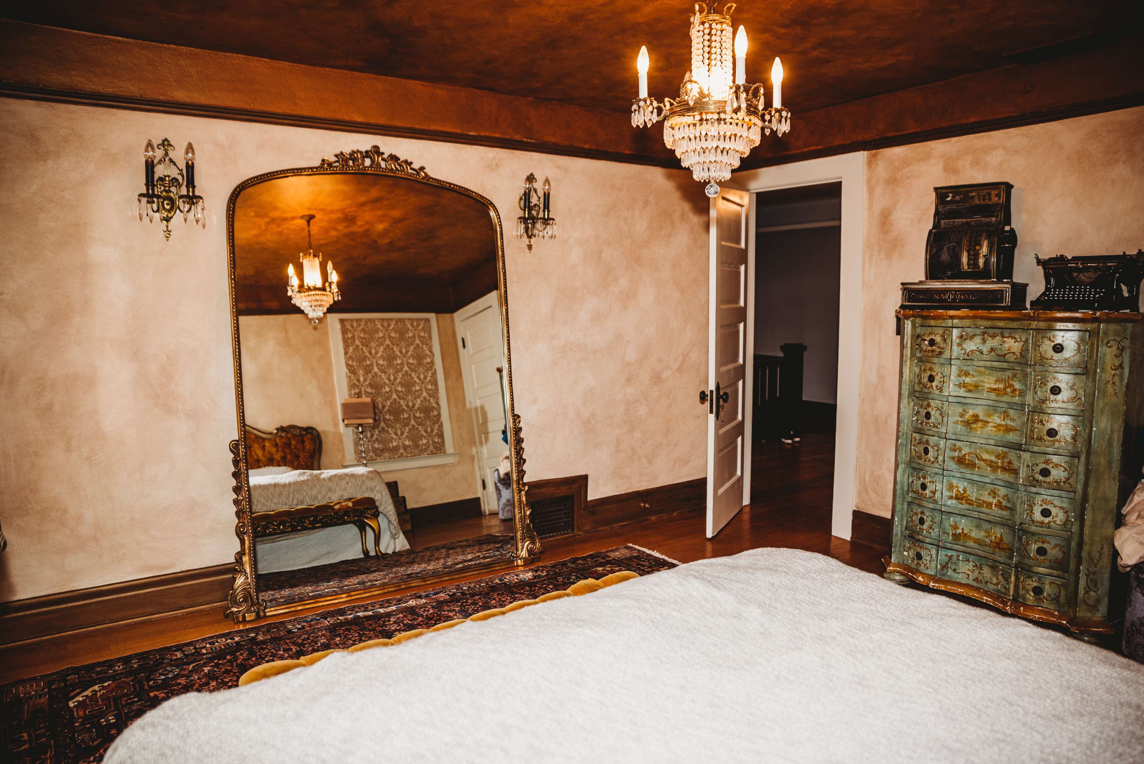 A vintage-style bedroom featuring a large ornate mirror, a bed with a light cover, a chandelier, and a decorative wooden cabinet against the wall.