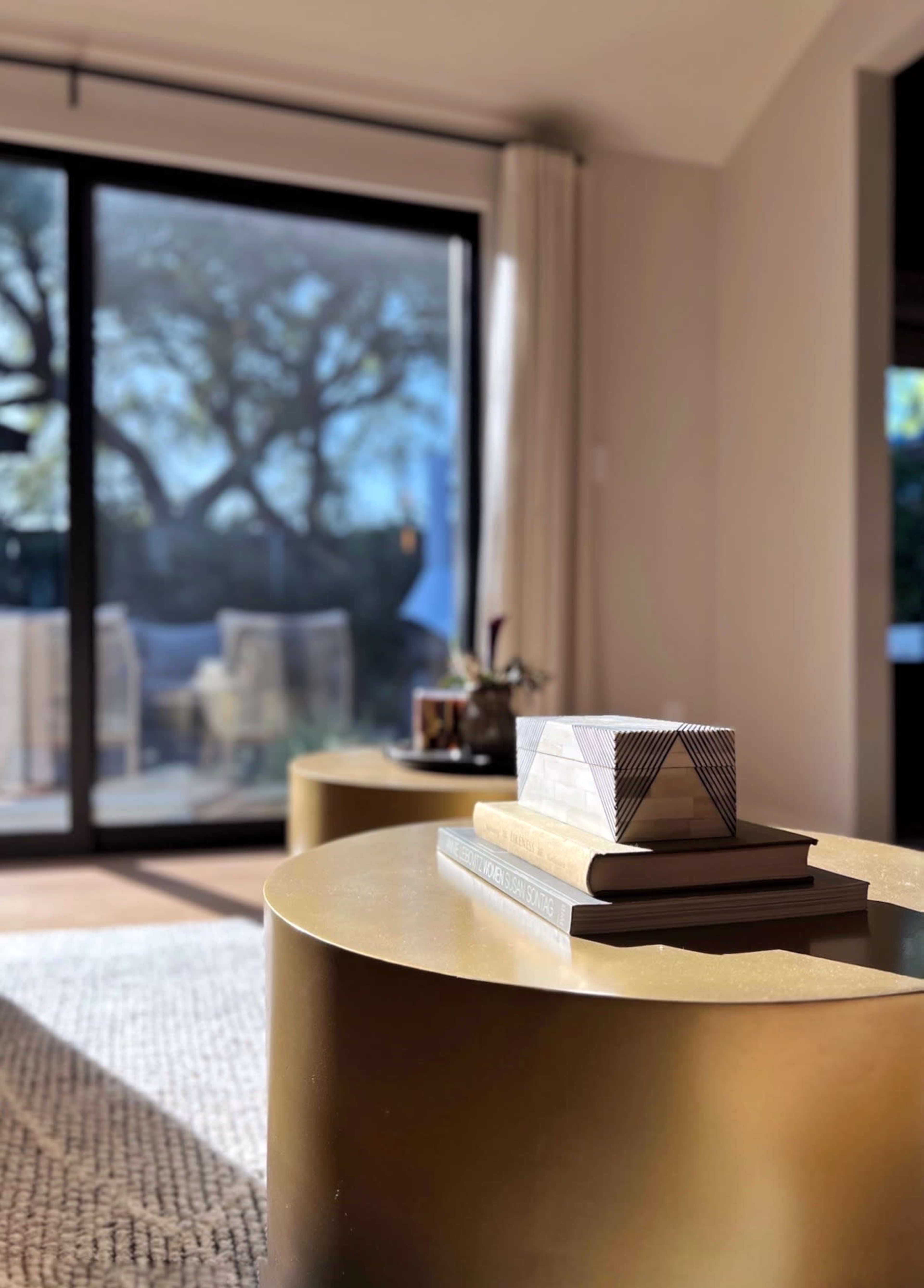 A geometric object rests on stacked books atop a gold coffee table in a well-lit living room with large glass doors leading to an outdoor space.