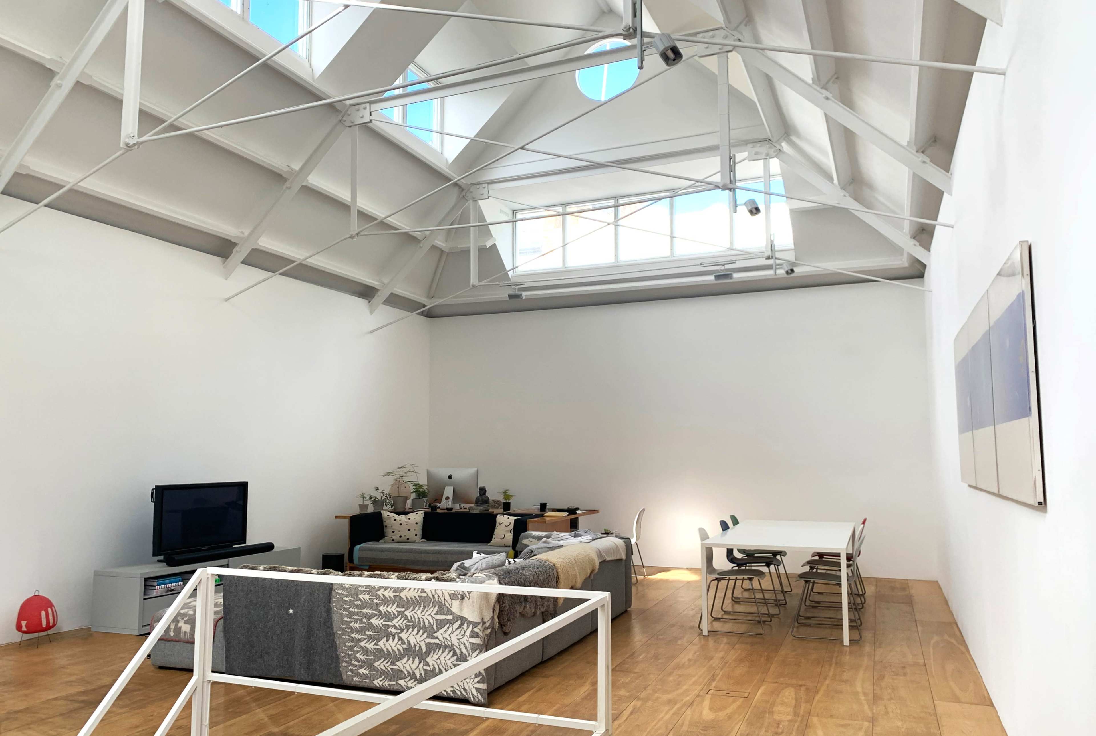 The image shows a spacious, modern room with a high ceiling, featuring a living area with a sofa and television, and a dining table with chairs.