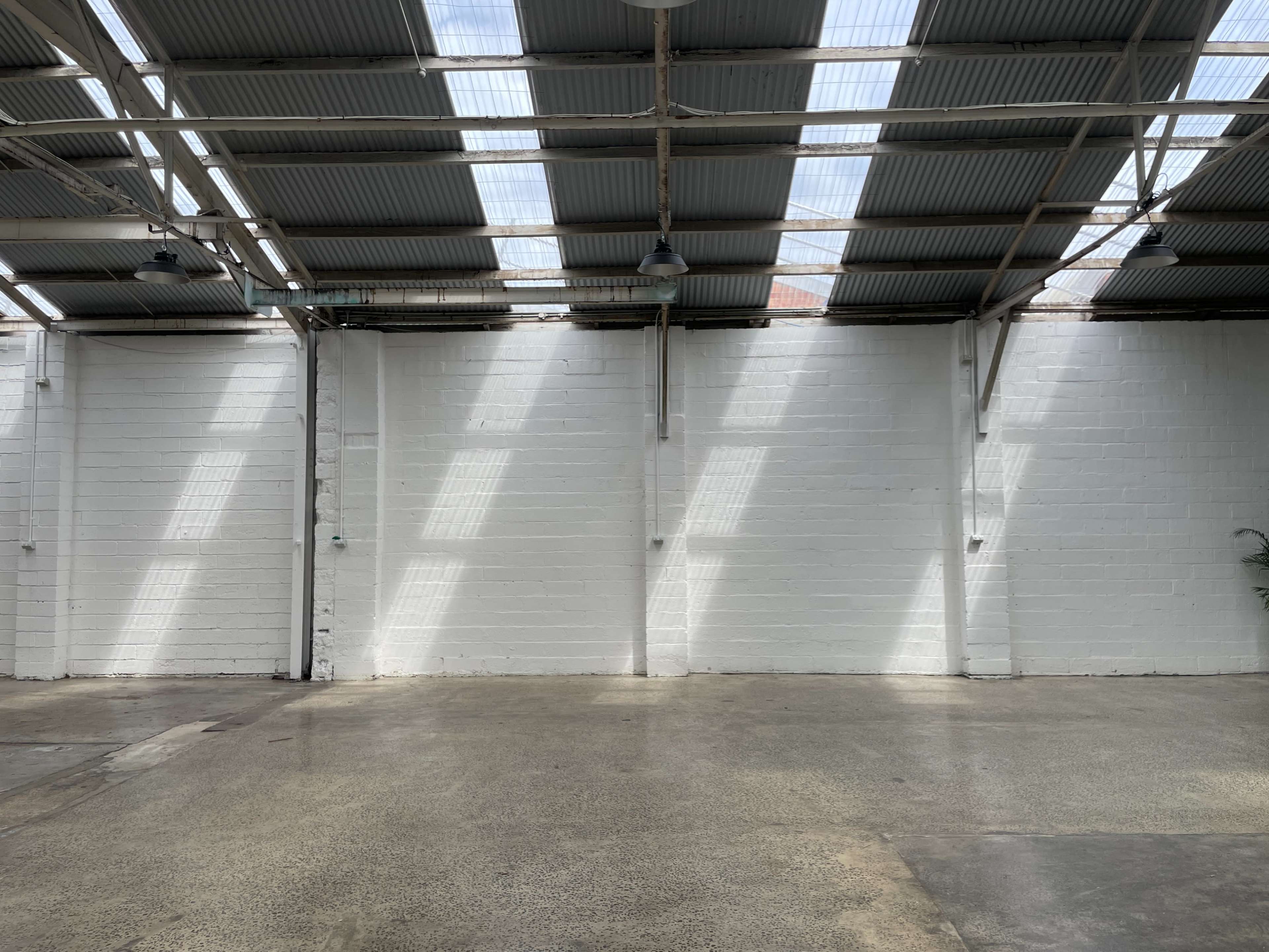 The image shows an empty warehouse with a concrete floor and sunlight streaming through skylights onto a white wall.