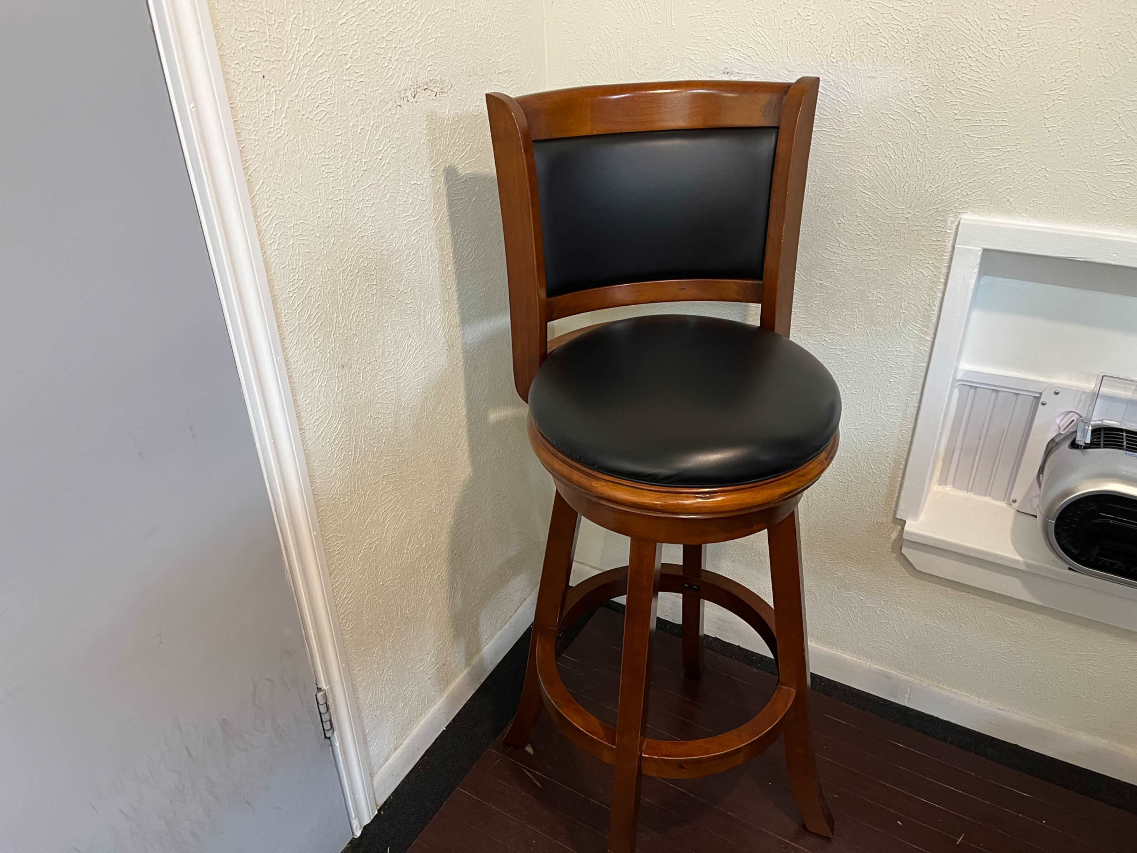 A wooden barstool with a black cushioned seat is positioned in a corner next to a wall.
