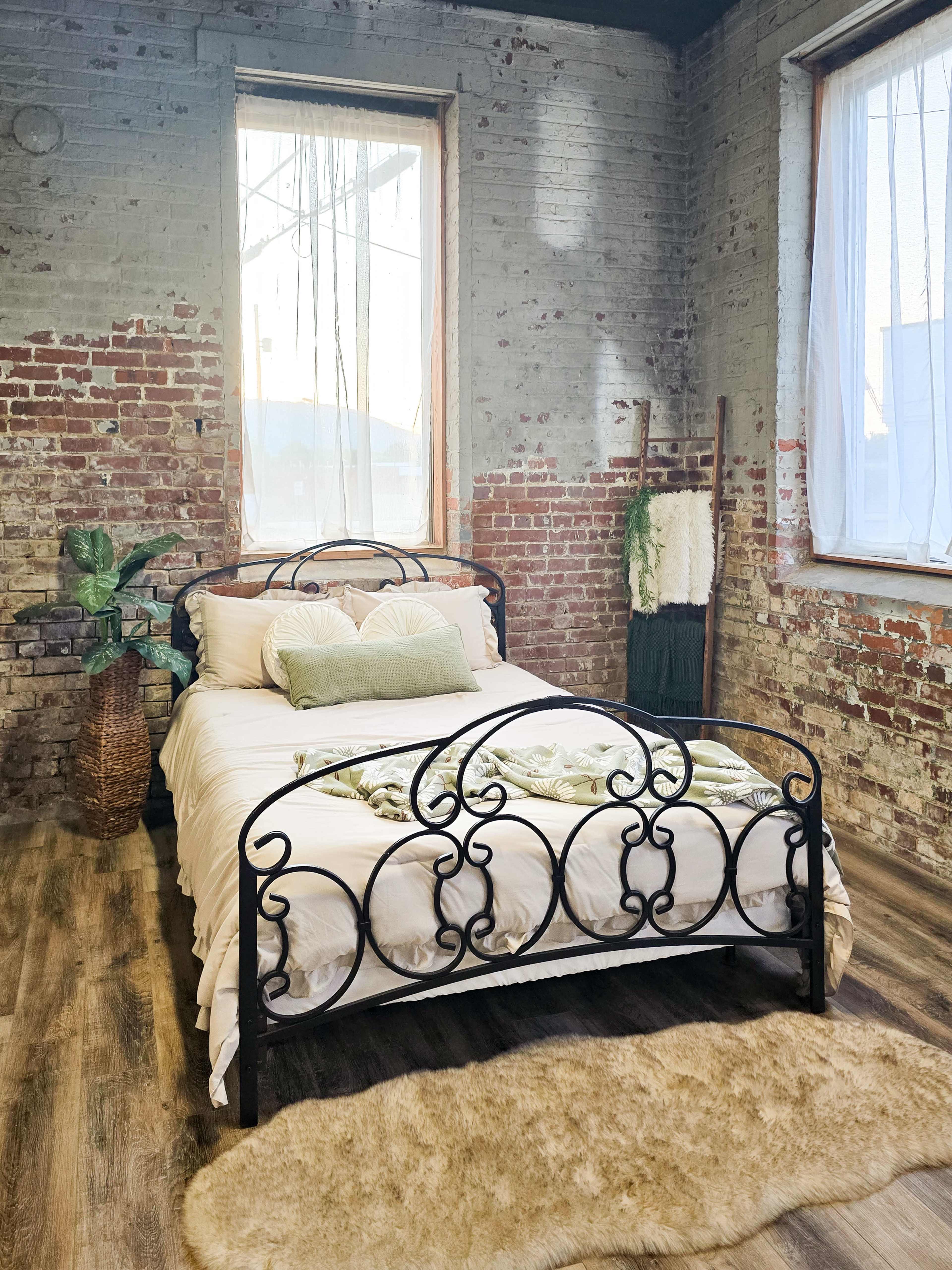 The image shows a bedroom featuring a metal bed frame with a cream-colored bedspread, two green decorative pillows, and a small area rug on a wooden floor, set against exposed brick walls and large windows.
