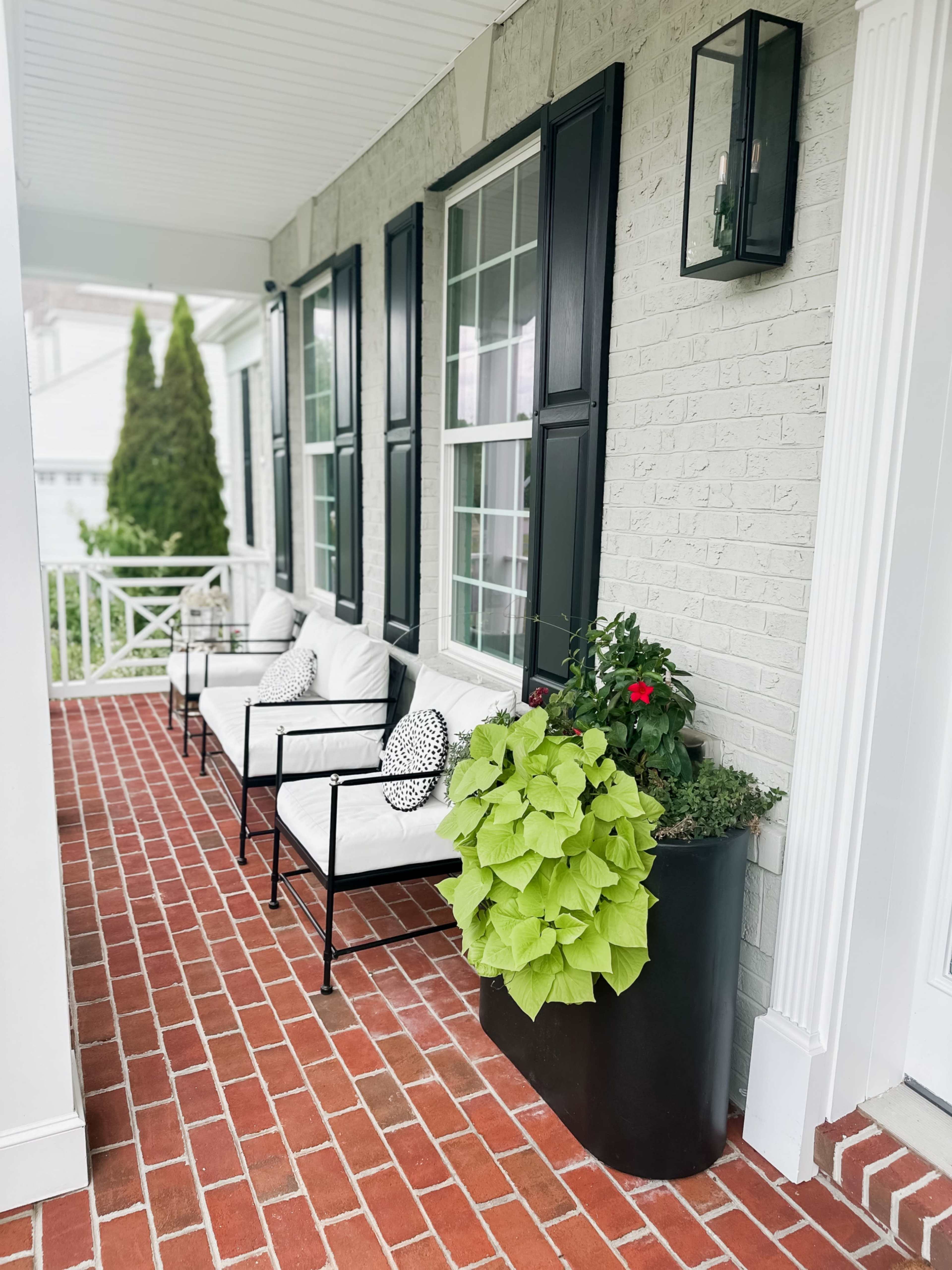 A porch with two black metal chairs and a planter filled with green plants and flowers, alongside a brick floor.