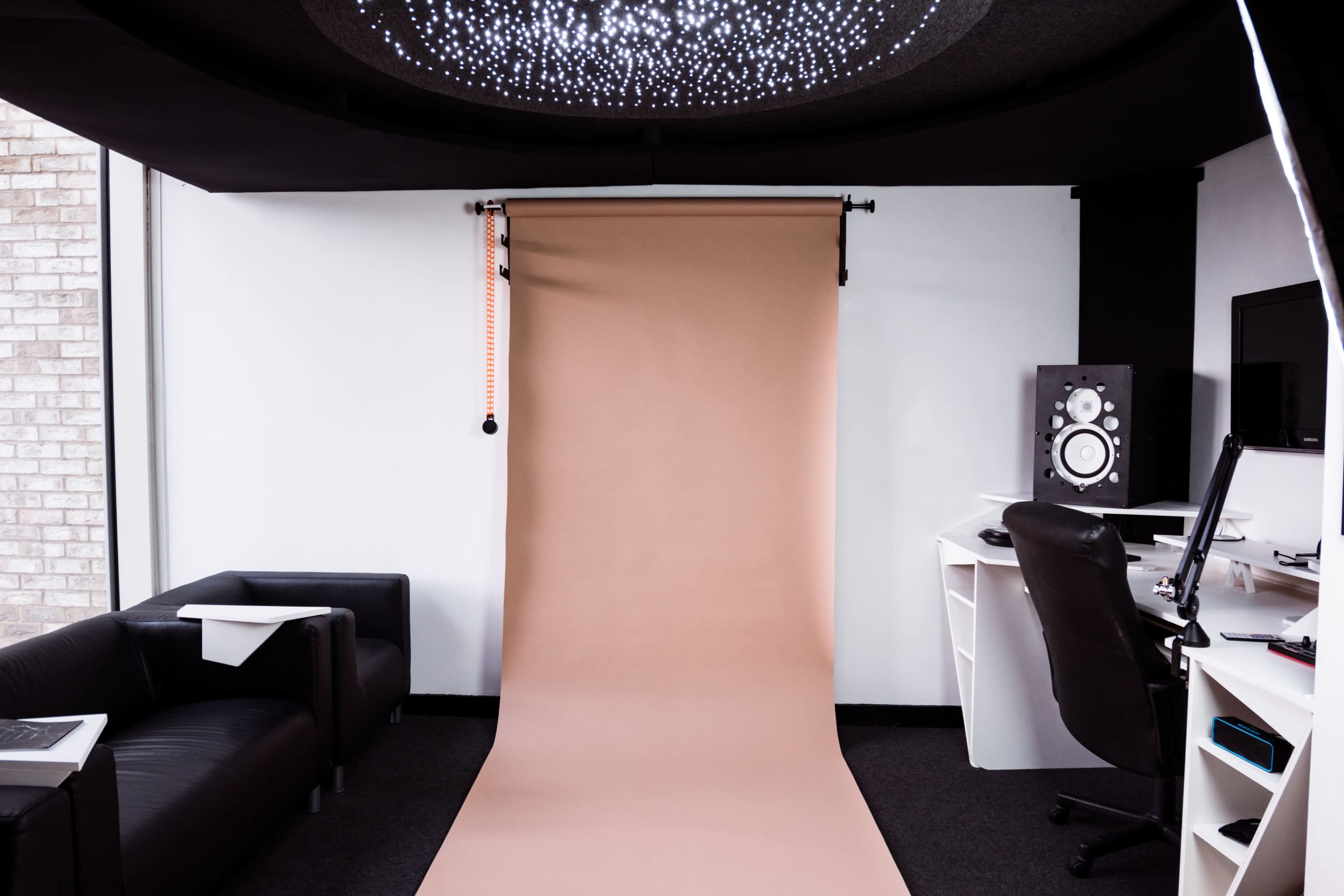 A photography studio with a beige backdrop, black leather seating, and a desk with audio equipment and a computer.