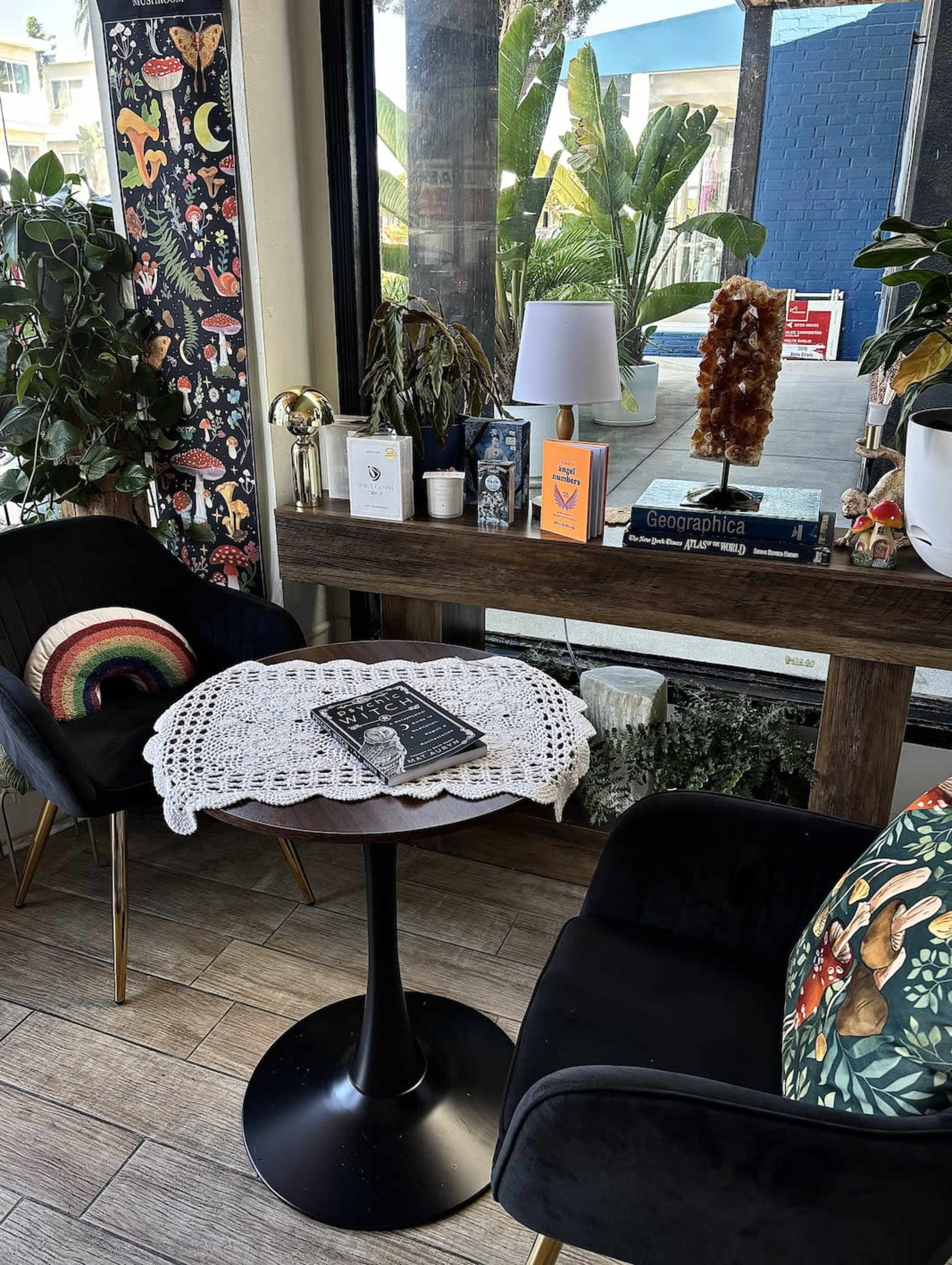 The image features a cozy seating area with two chairs, a small round table topped with a lace doily and books, and decor elements like plants and artworks in a well-lit environment.