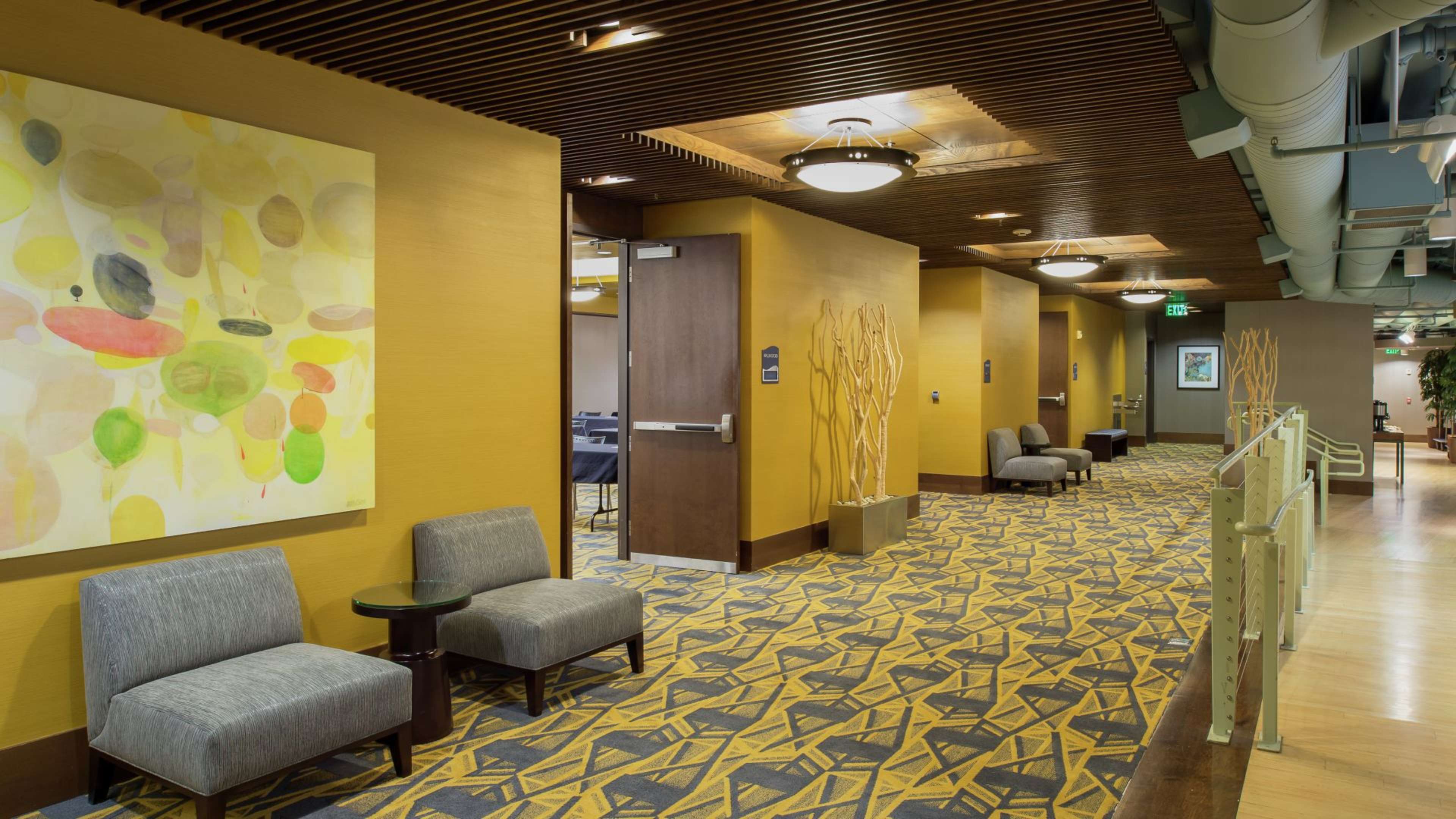 A corridor with yellow walls, modern artwork, and a patterned carpet, featuring seating areas along the sides.