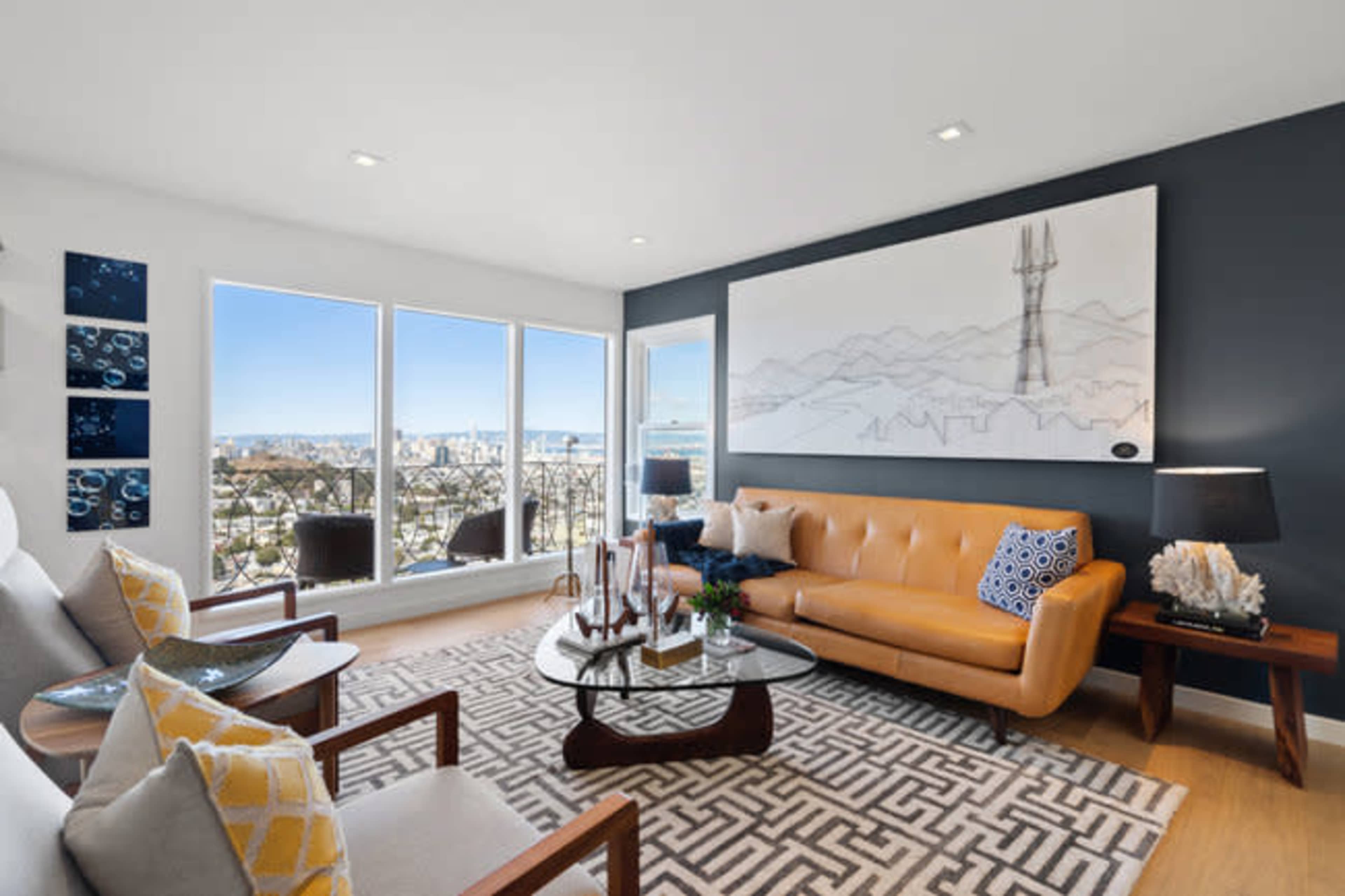 A modern living room with a large window overlooking a cityscape, featuring a brown leather sofa, two armchairs, and a large wall art piece.