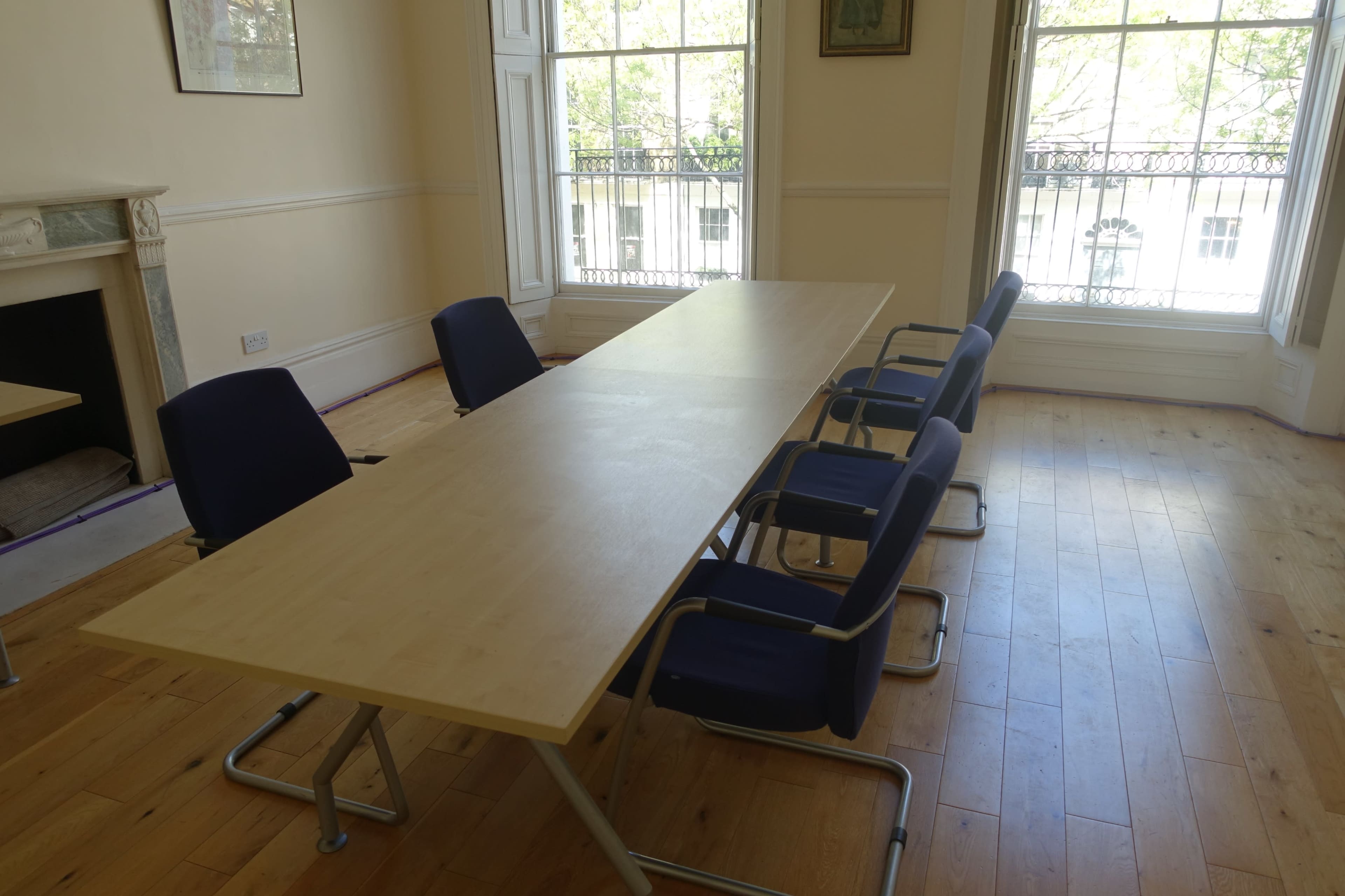 A large rectangular table with several blue chairs surrounds it in a sunlit meeting room with large windows.