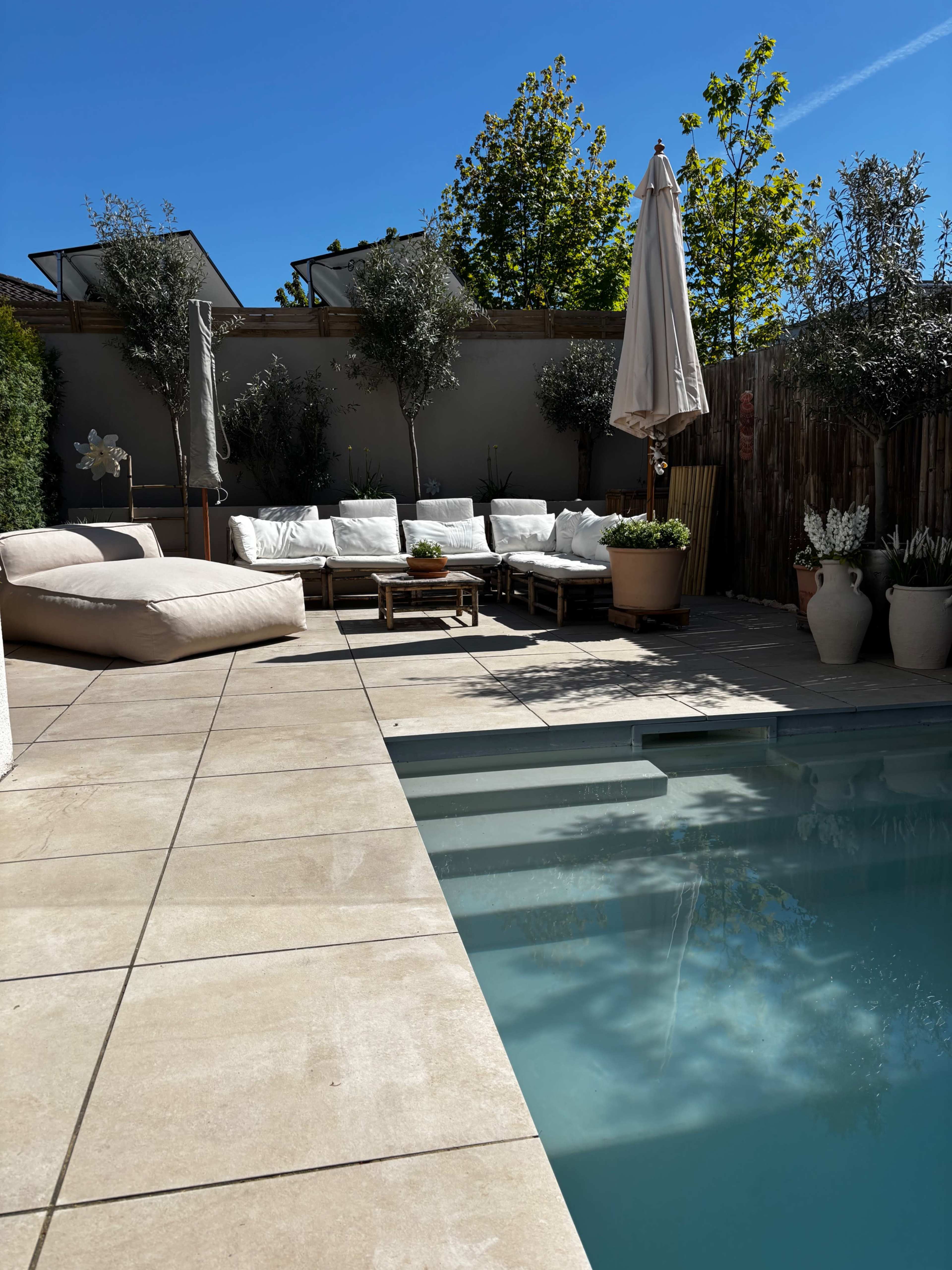 The image shows a patio area with a swimming pool, sun loungers, and a seating arrangement surrounded by potted plants under clear blue skies.