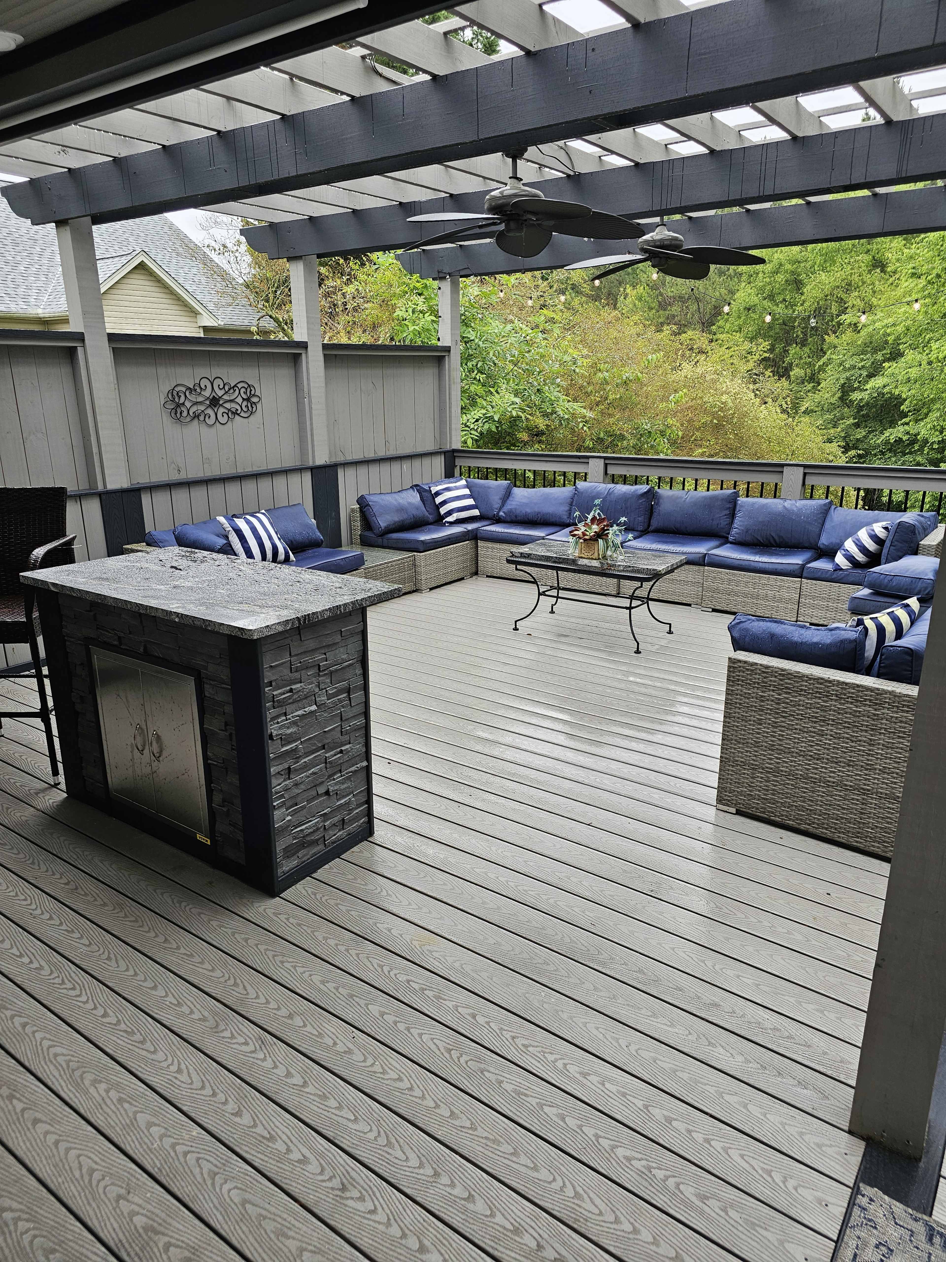 A patio area features a seating arrangement with blue cushions, a coffee table, and a stone outdoor bar under a slatted pergola.