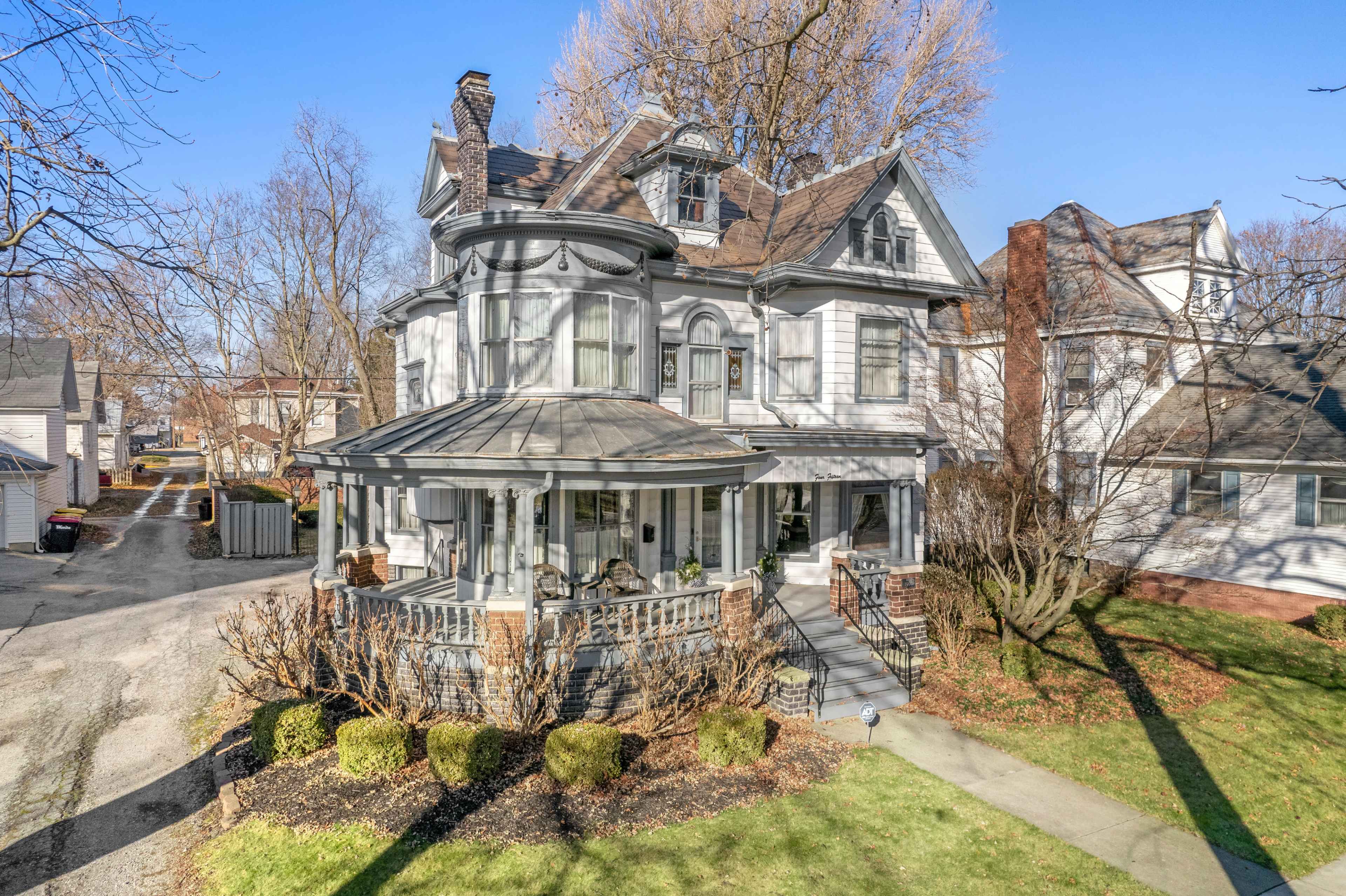 A large, two-story Victorian-style house with a wraparound porch and a conical turret, surrounded by manicured landscaping in a residential neighborhood.