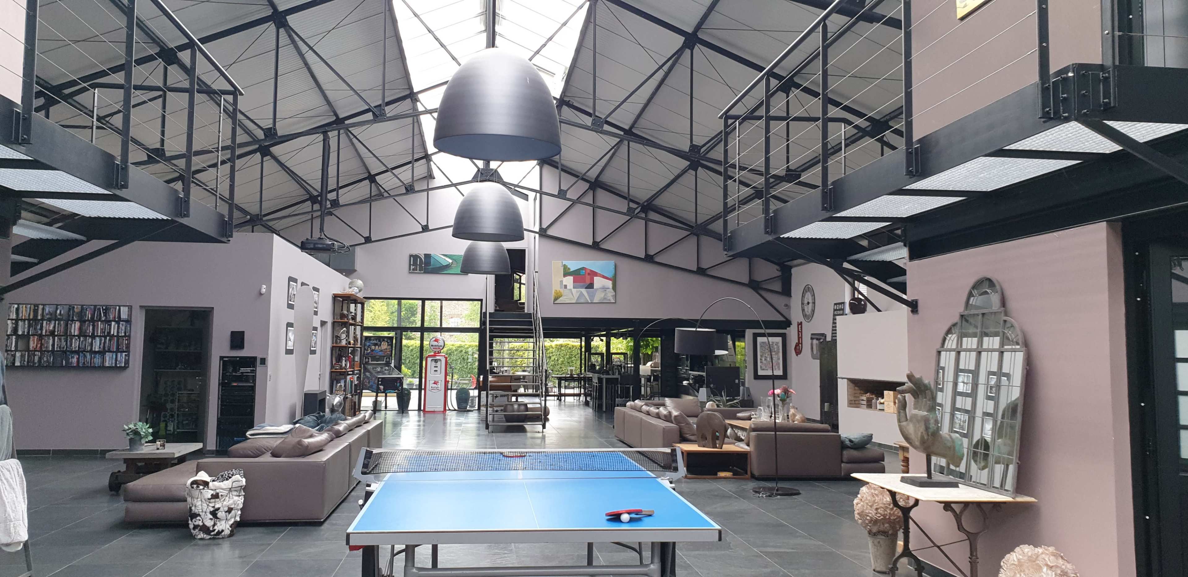 A spacious, modern indoor area with a blue ping-pong table at the center, surrounded by seating arrangements and large windows.