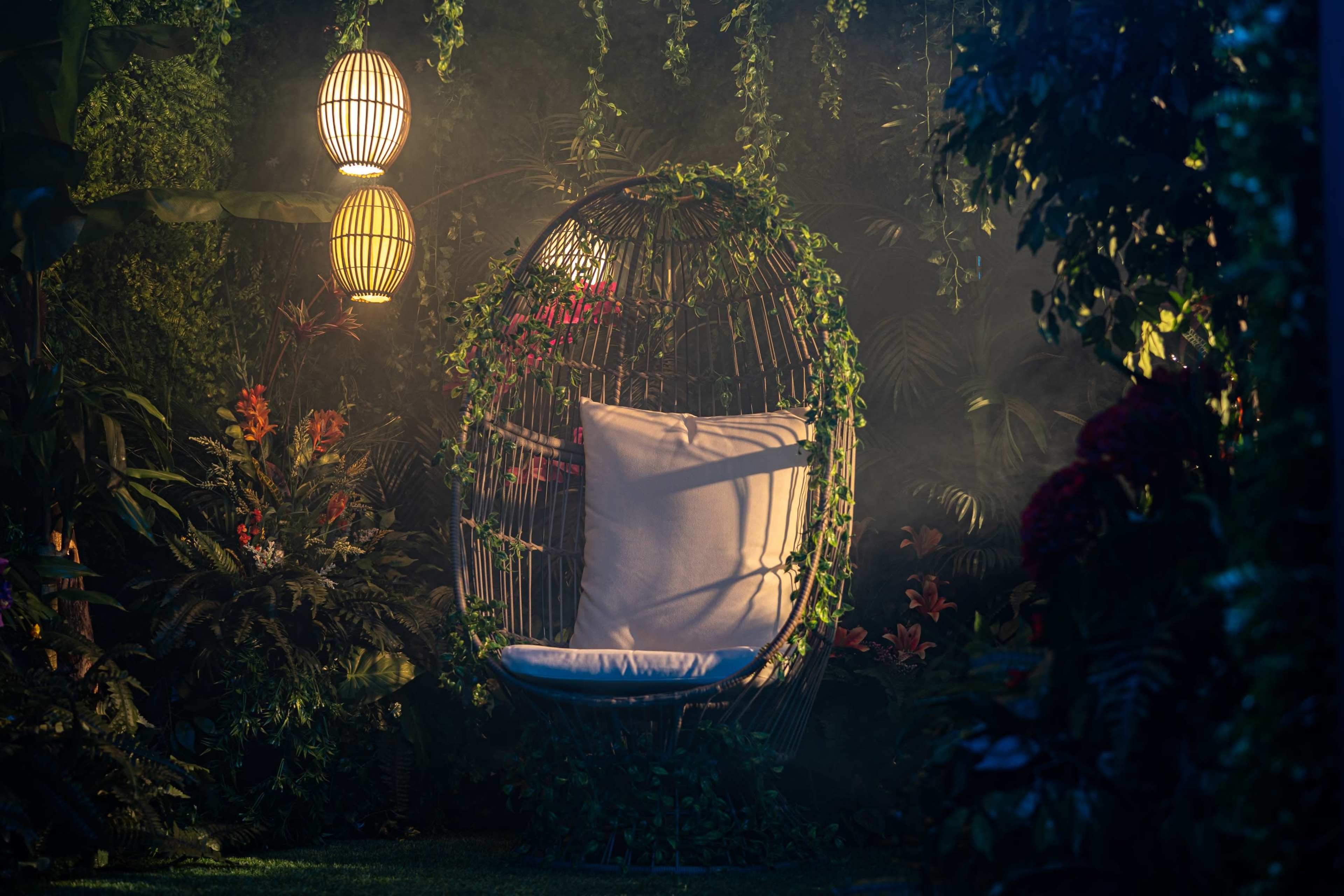 A suspended, wicker egg chair with white cushions is surrounded by lush greenery and illuminated by ambient lanterns.
