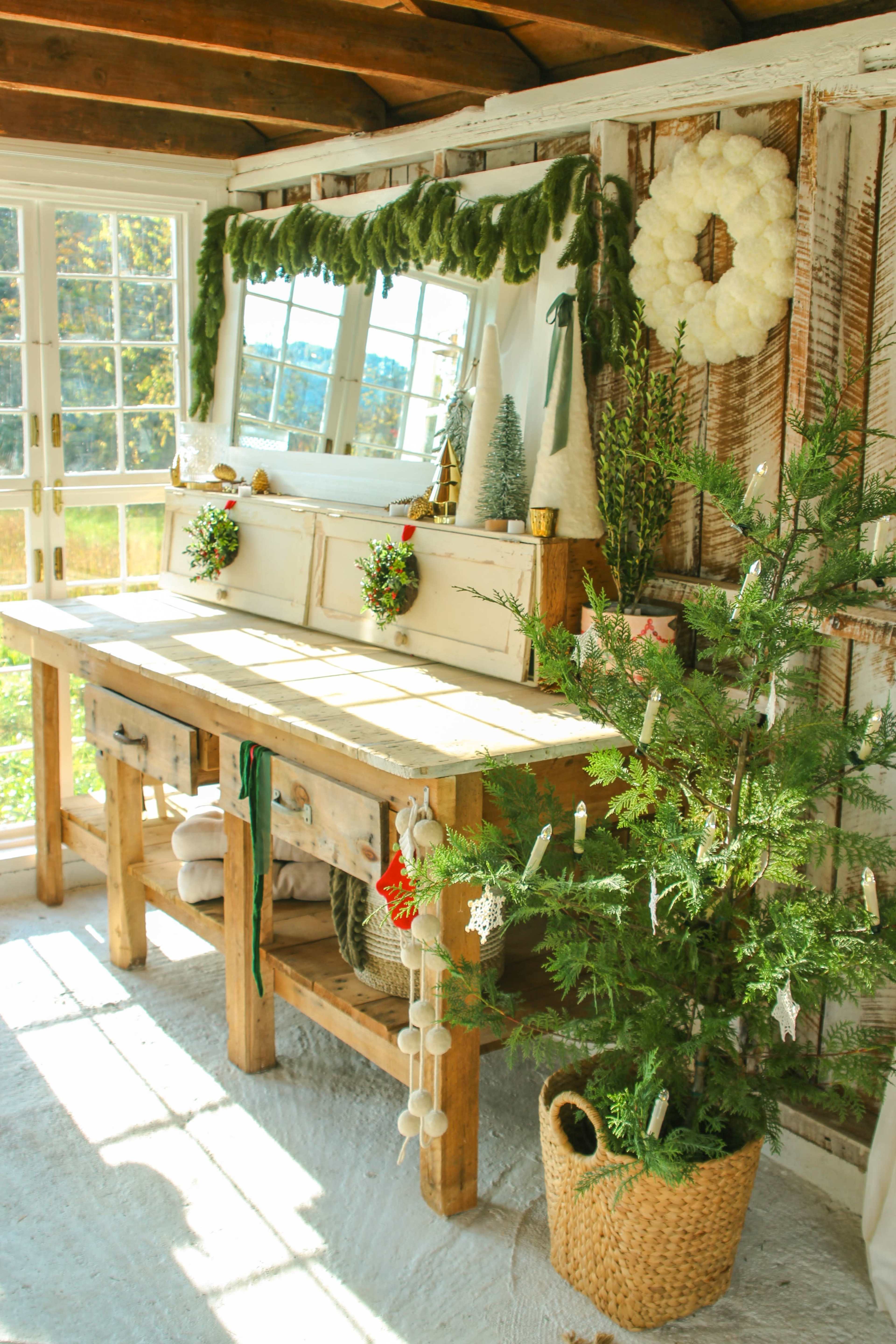 A rustic wooden table is set against a wall with large windows, decorated with a mirror, greenery, and holiday ornaments, alongside a small Christmas tree in a woven basket.