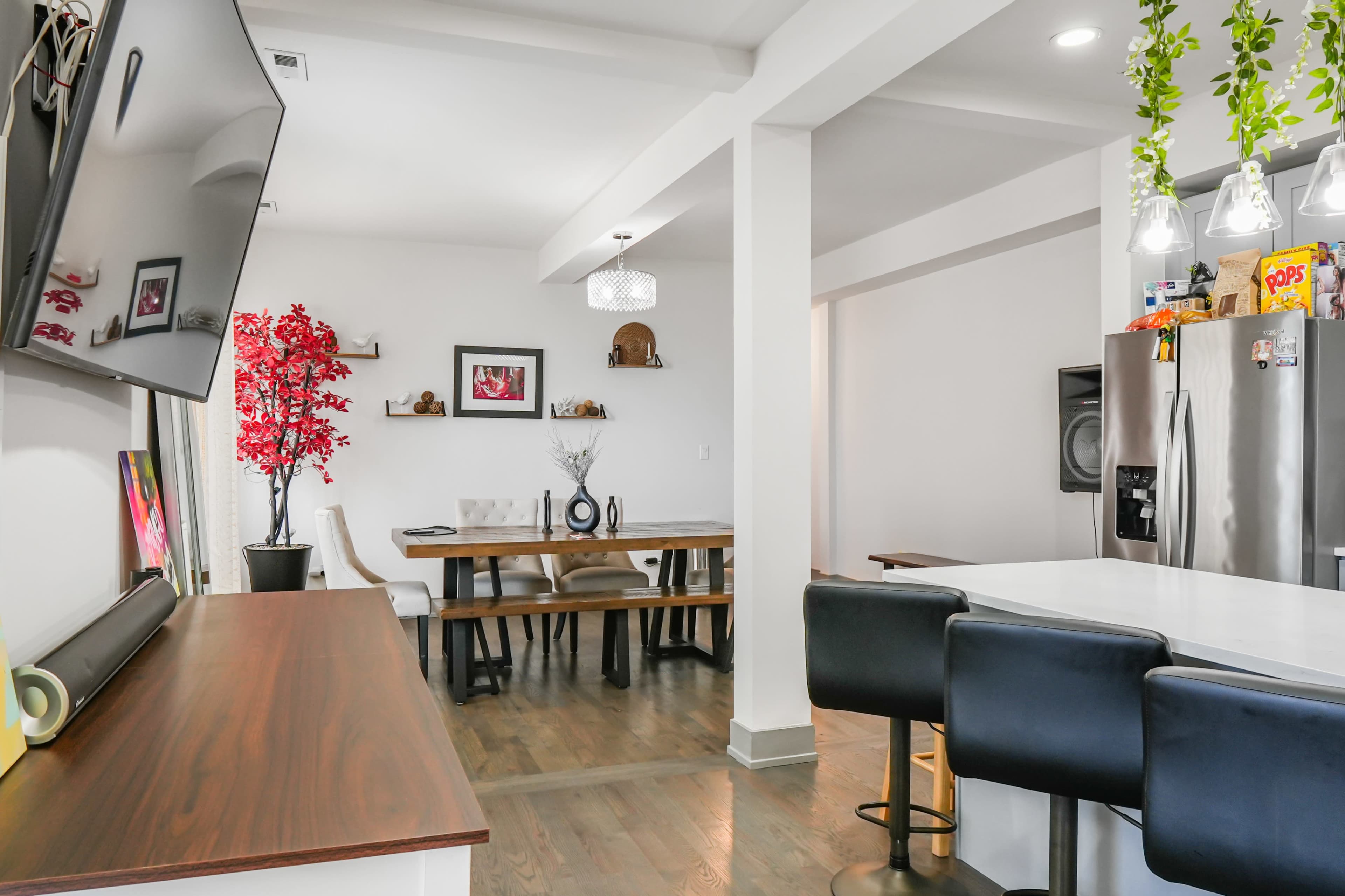 The image shows a modern kitchen and dining area featuring a dining table, bar seating, and decorative plants.