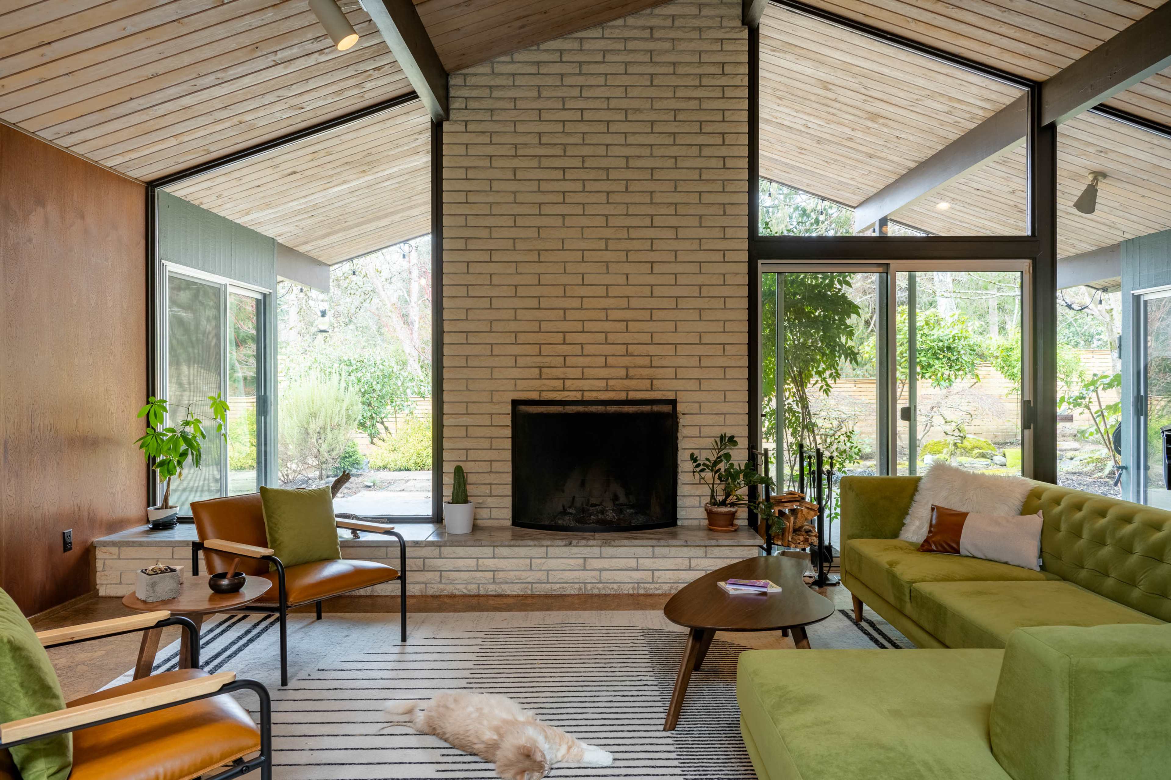 A modern living room features a stone fireplace, large windows with views of greenery, and several seating options including a green sofa and brown chairs.