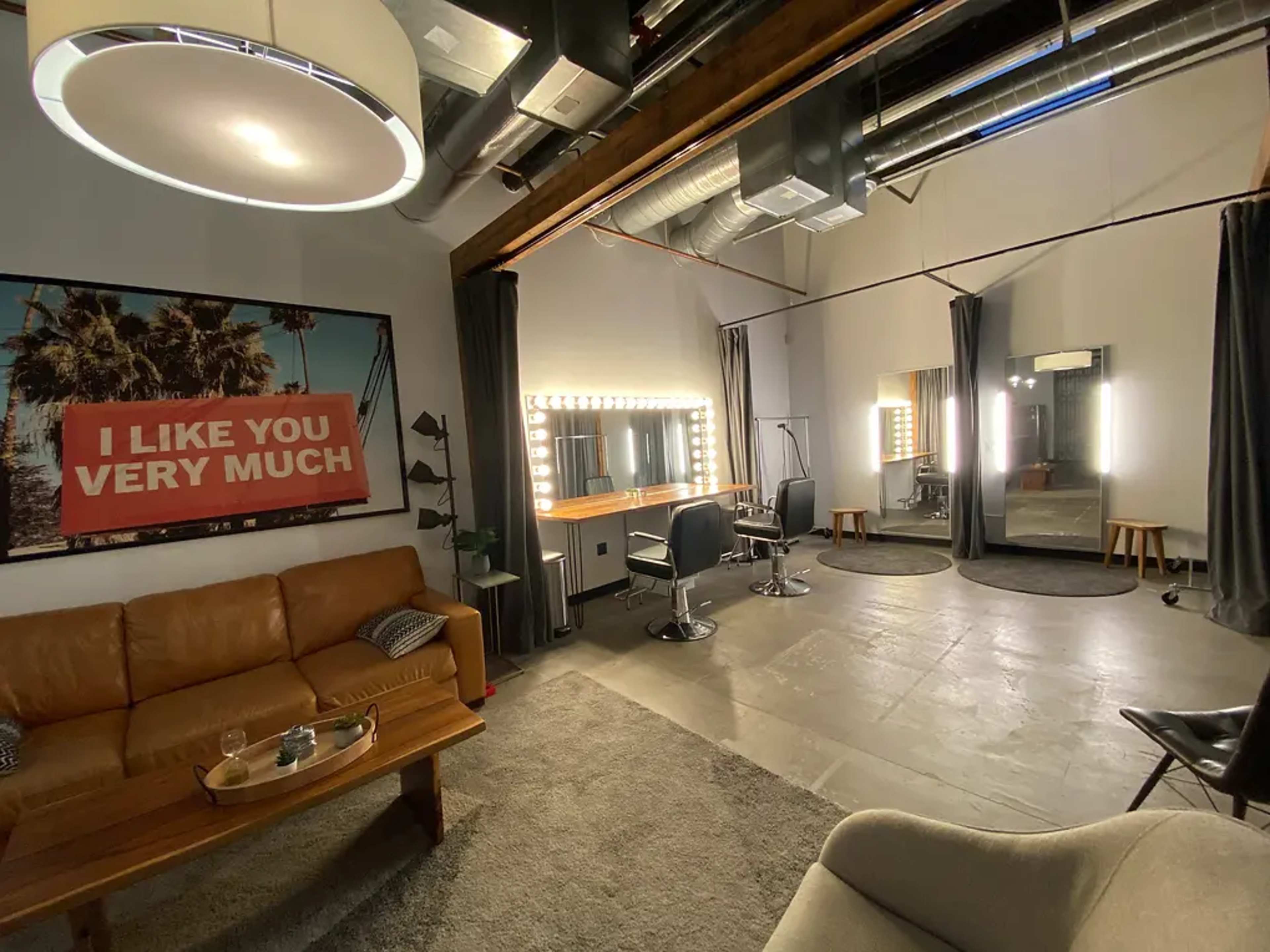 The image shows a modern, spacious studio room featuring a leather sofa, a large mirror with lights, and several chairs, along with decorative curtains and a wall art piece.