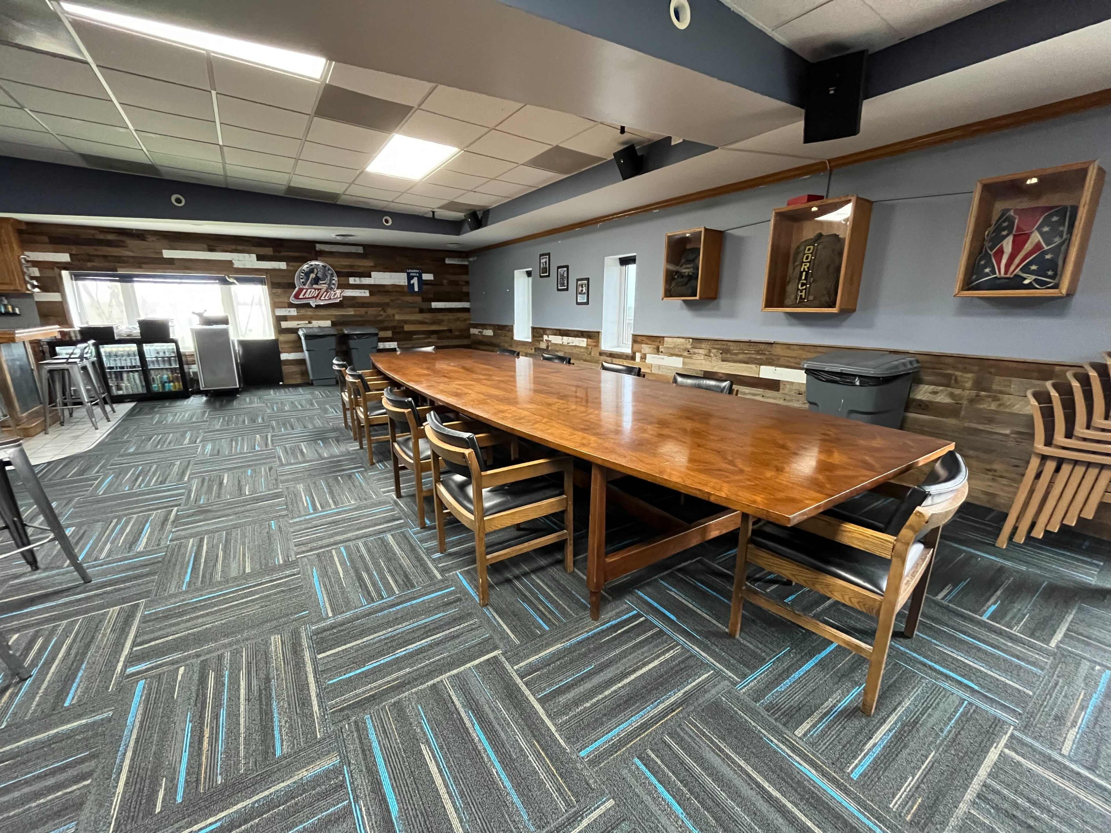 The image shows a spacious meeting room with a large wooden table surrounded by chairs, featuring a patterned carpet and wooden wall accents.