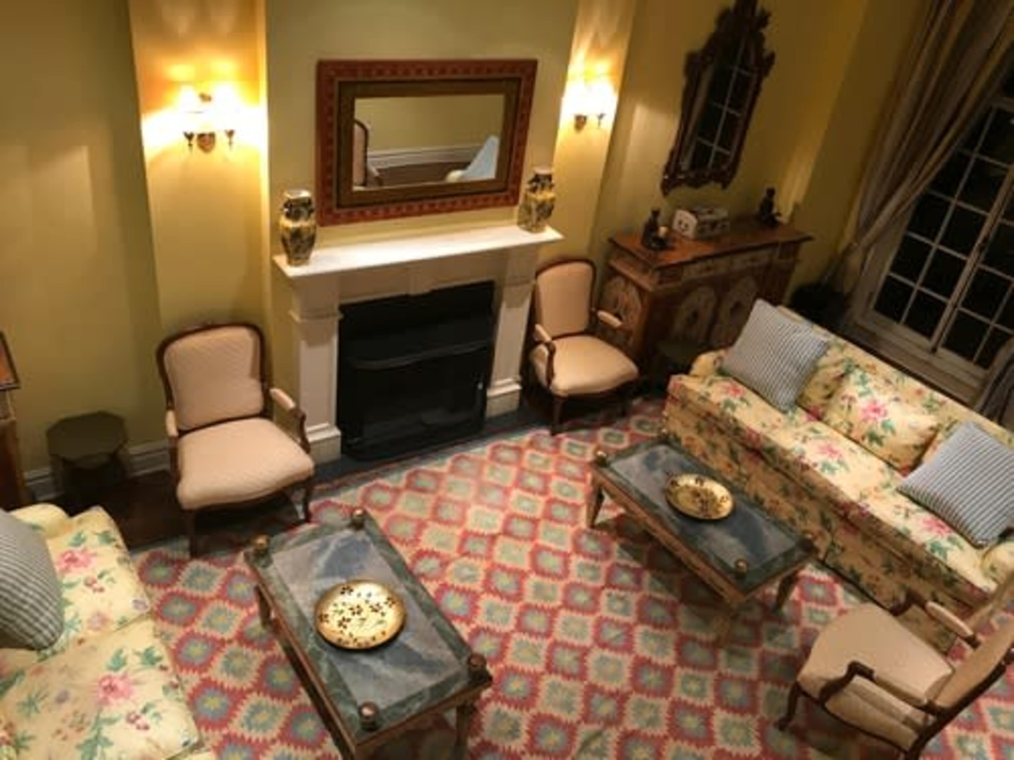 A cozy living room with floral-patterned sofas, two armchairs, a fireplace, a decorative mirror, and a patterned rug.