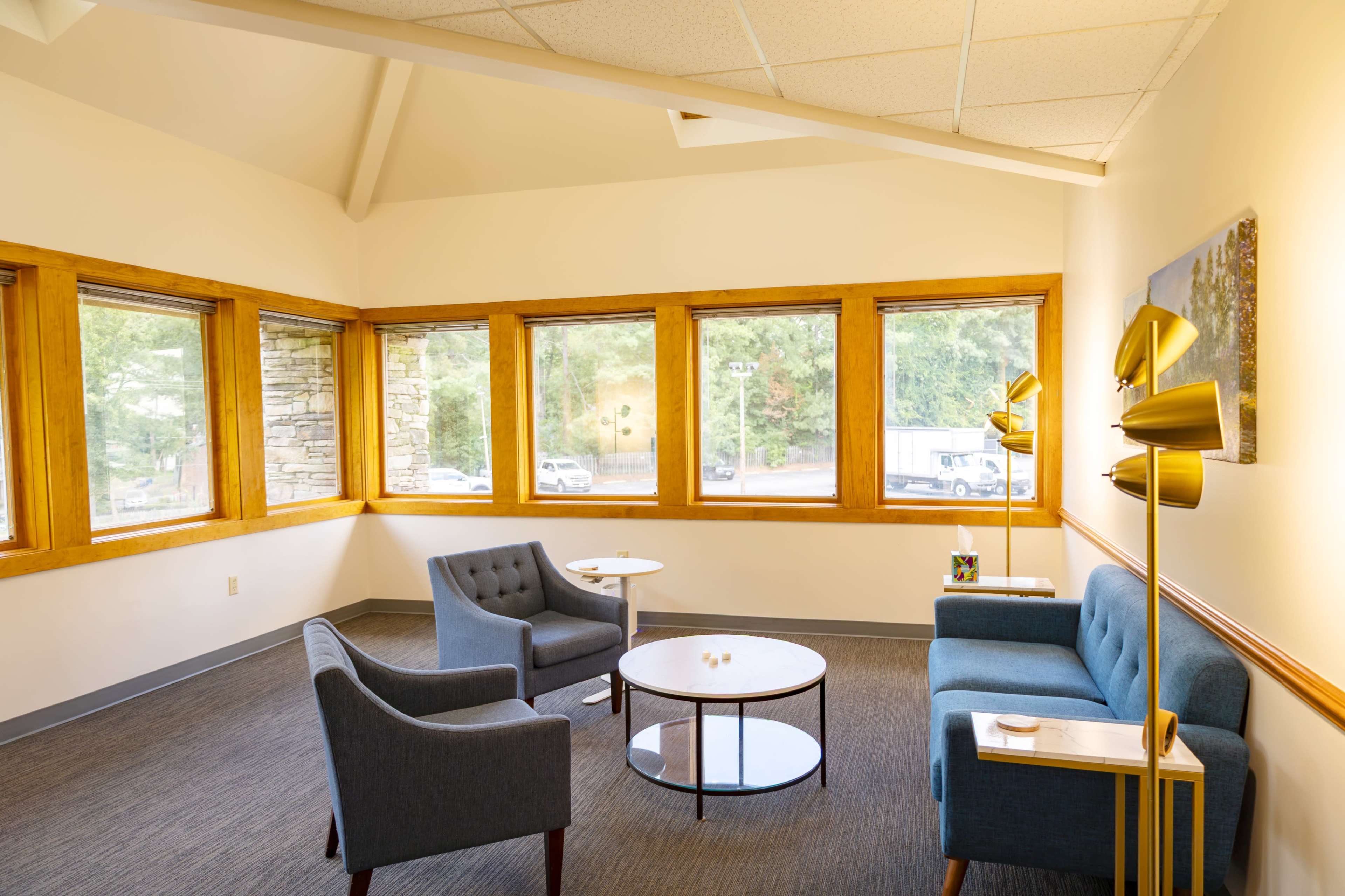 The image shows a bright, modern waiting area with two blue couches, a round coffee table, and multiple large windows allowing natural light to enter.