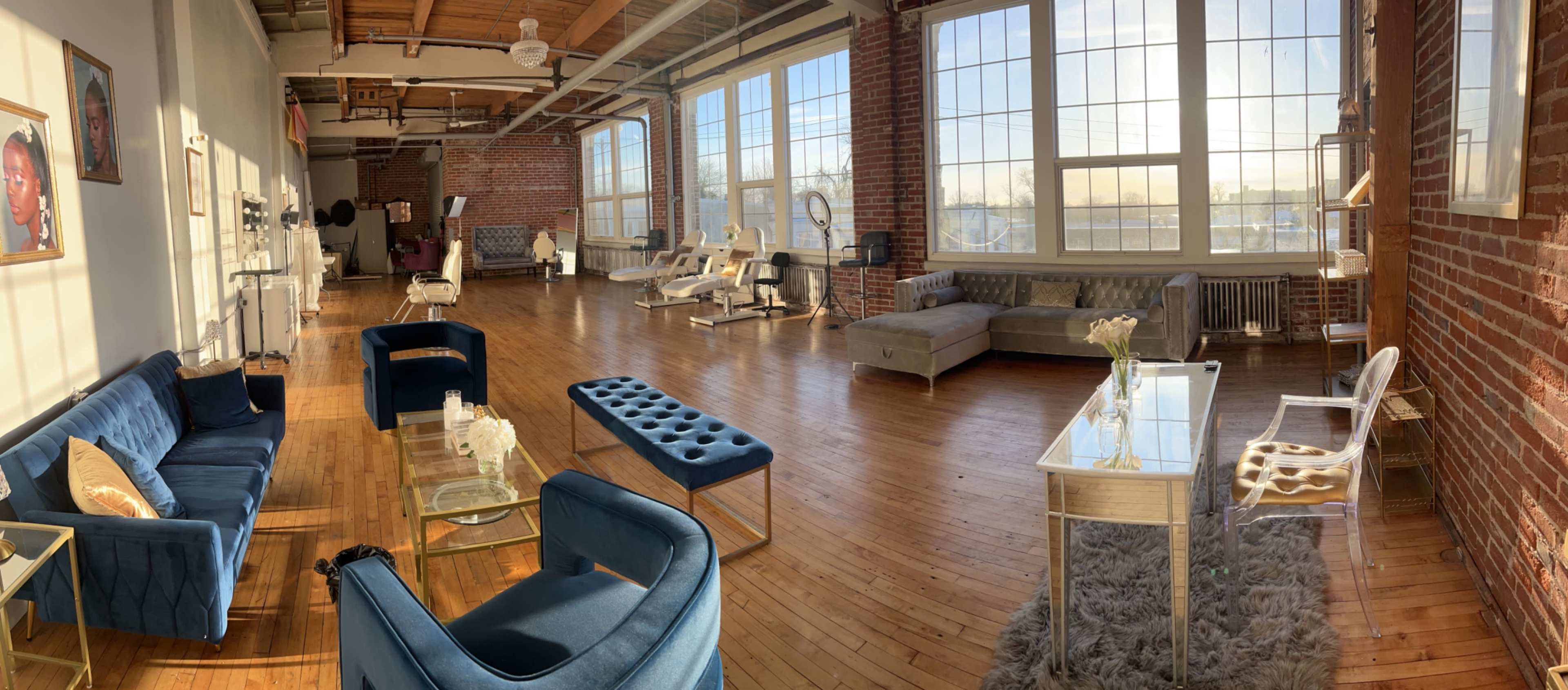 The image shows a spacious, well-lit room with large windows, wooden flooring, and several seating areas, including sofas and chairs, accompanied by beauty treatment stations.