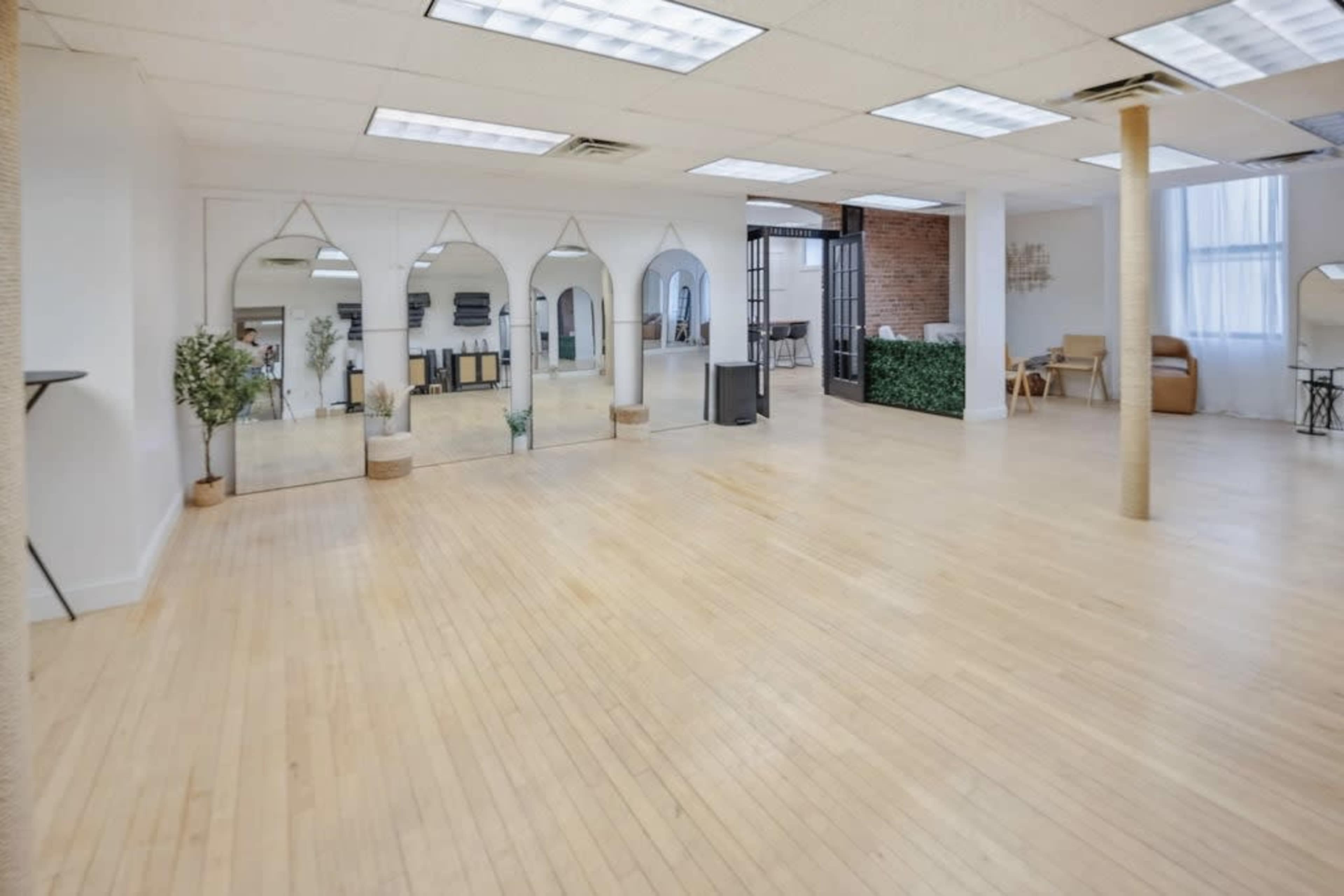 The image shows a spacious, empty room with wooden flooring, large mirrors, and natural light coming through the windows.