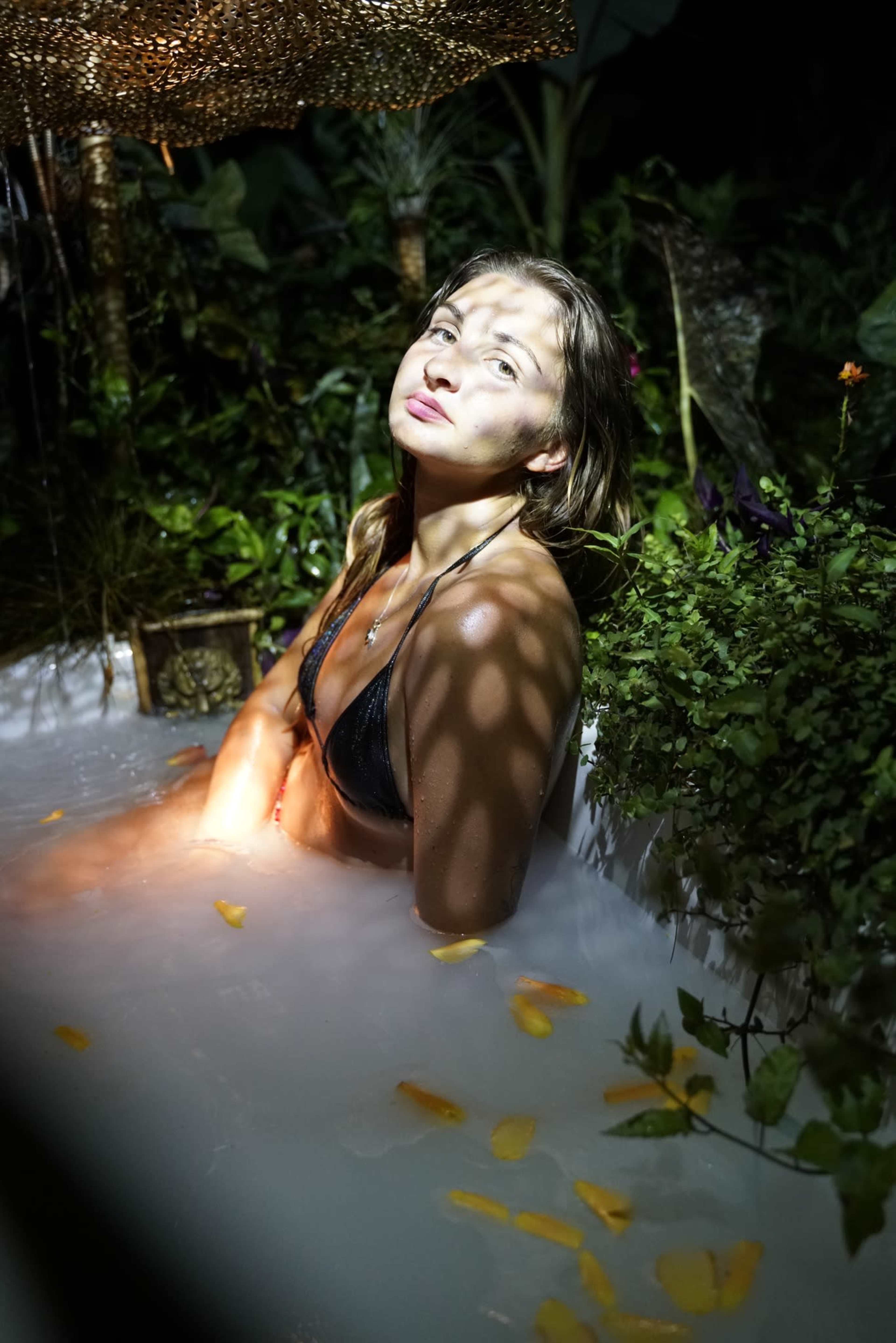 A woman with wet hair sits in a spa-like setting filled with mist, surrounded by foliage and illuminated by soft lighting.