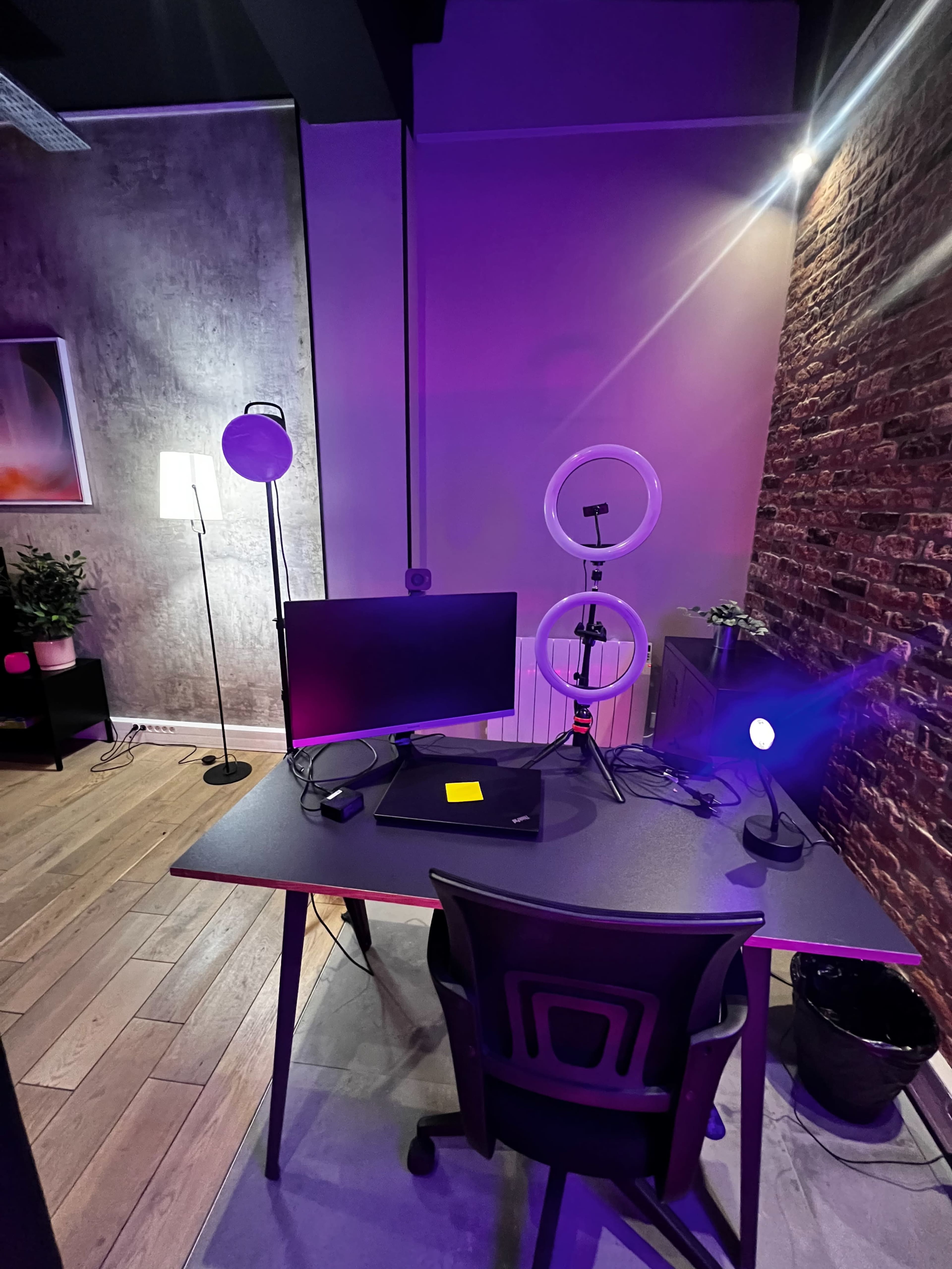 A modern workspace features a desk with a monitor and laptop, surrounded by decorative lighting and a plant.