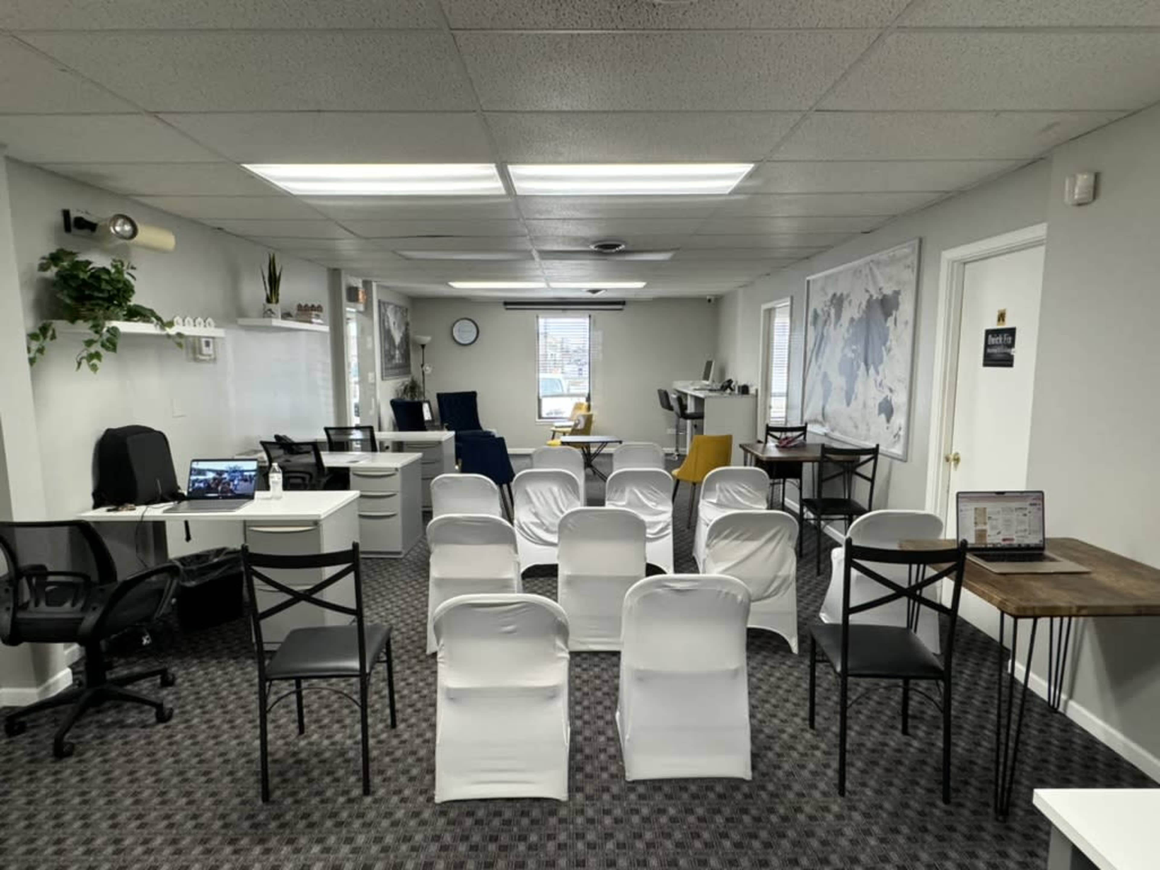 A minimalist office space is arranged with white chairs in rows, flanked by desks and computers, and featuring large windows and a wall map.