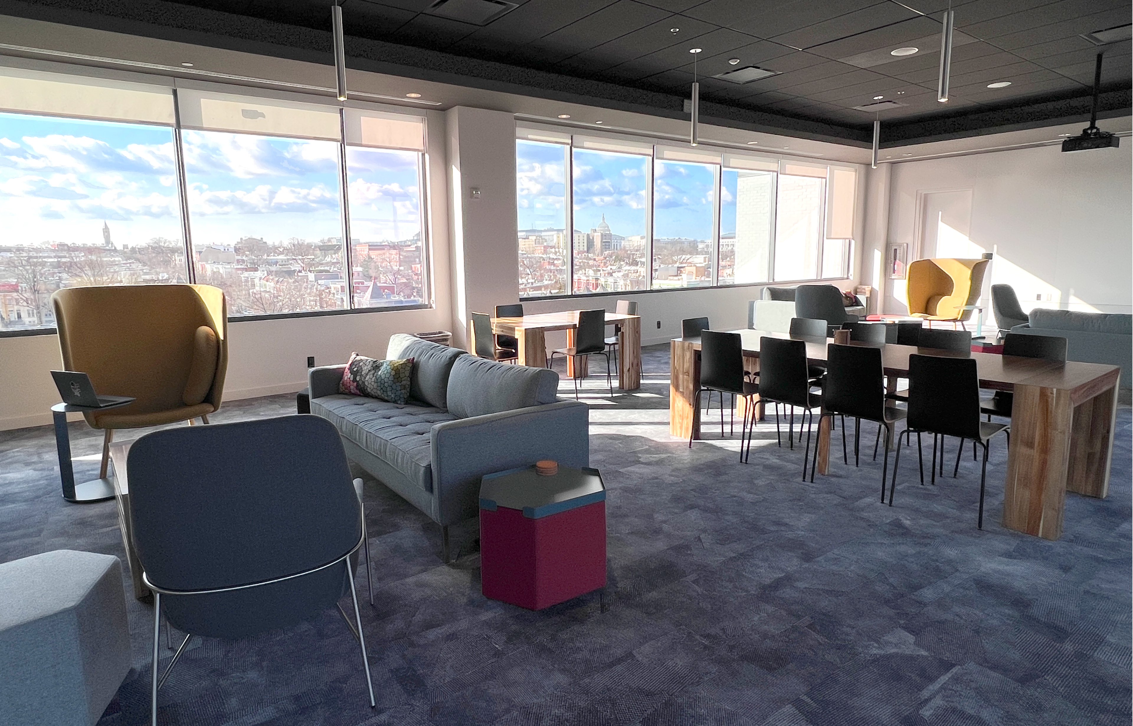 A modern office space featuring a seating area with a couch and chairs, a dining table, and large windows overlooking a city skyline.