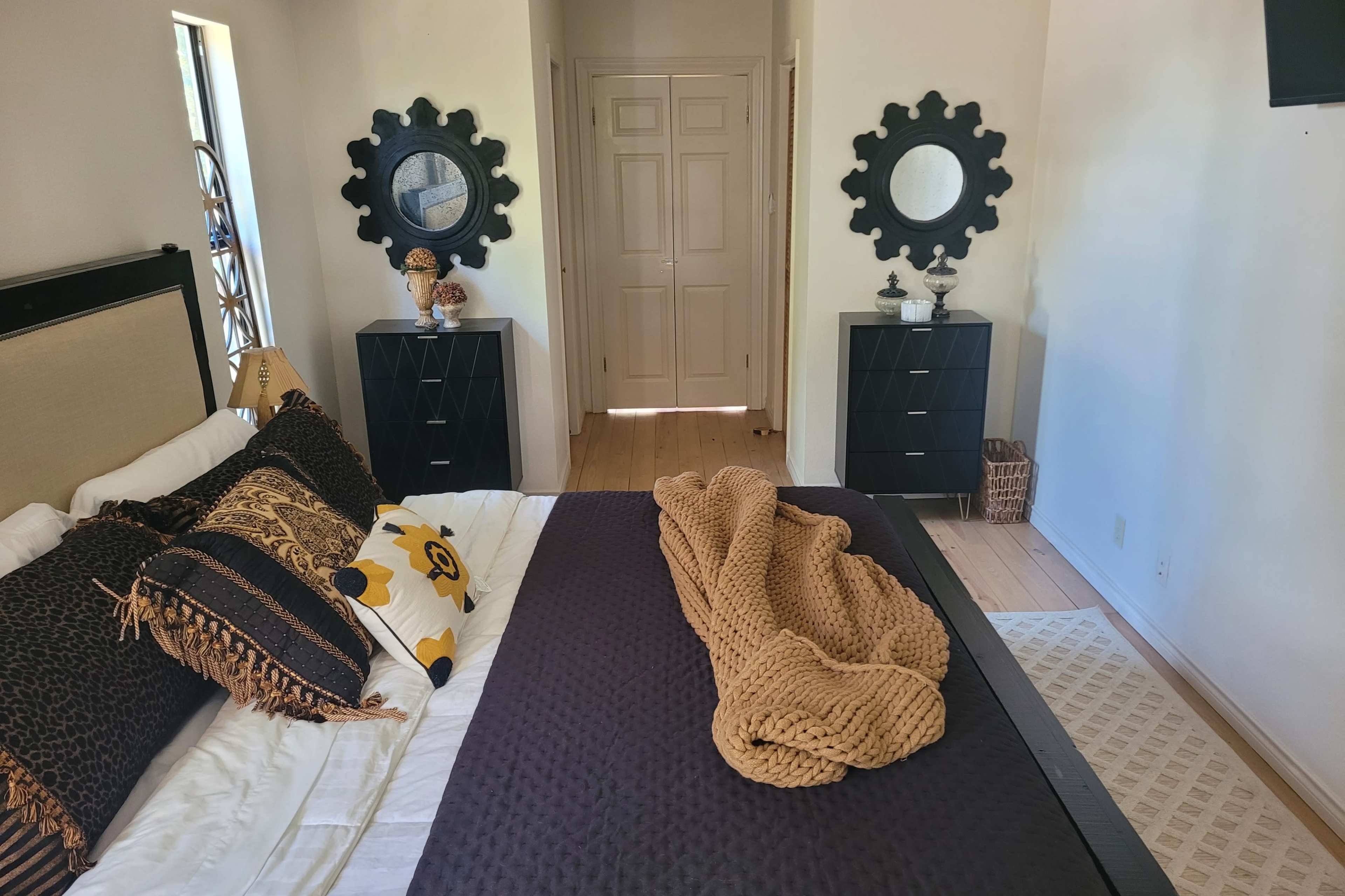 The image shows a neatly arranged bedroom featuring a bed with decorative pillows and a knitted blanket, flanked by two dark wooden dressers and mirrors on the walls.
