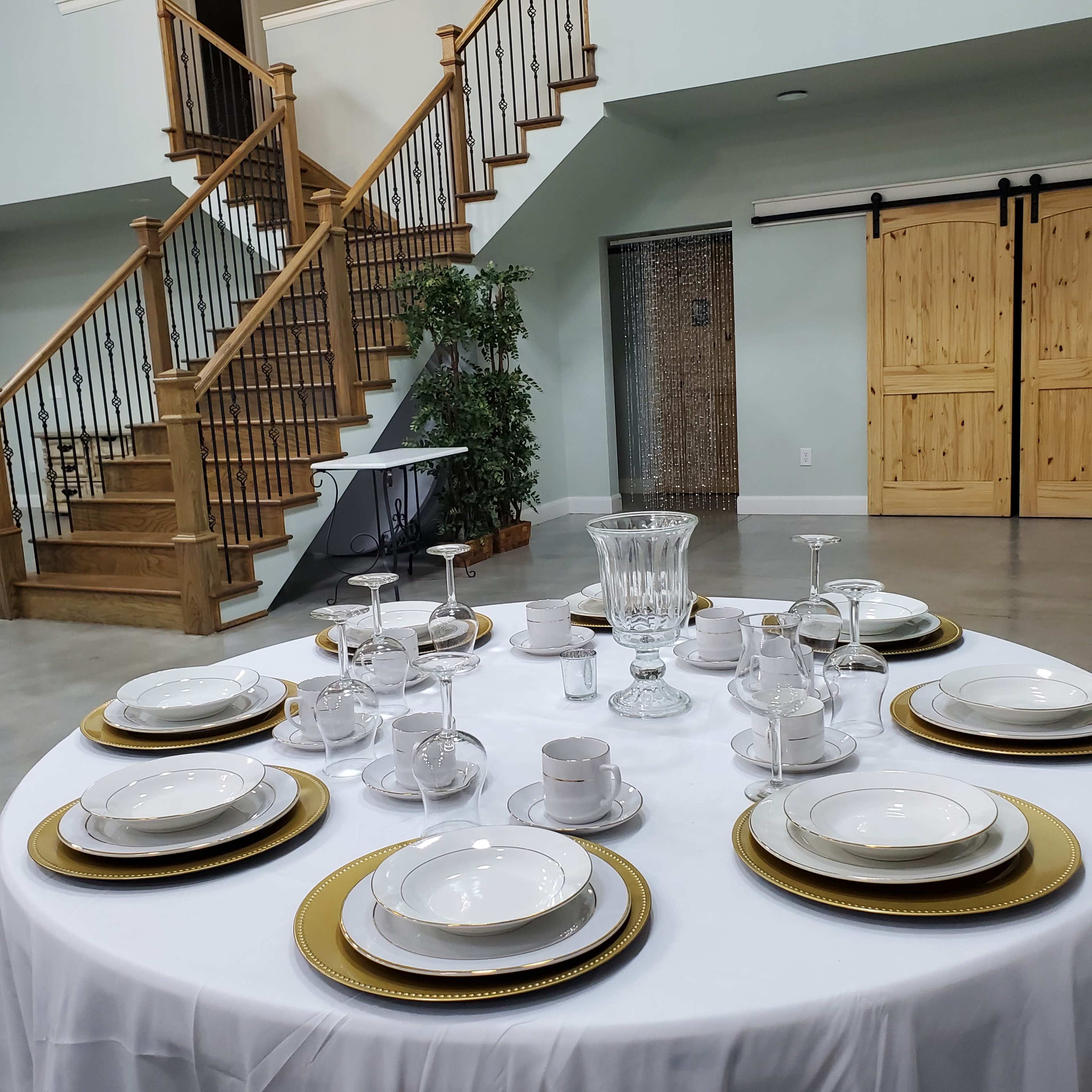 A round dining table is set with elegant dinnerware and glassware, surrounded by a staircase and decorative elements in a spacious room.