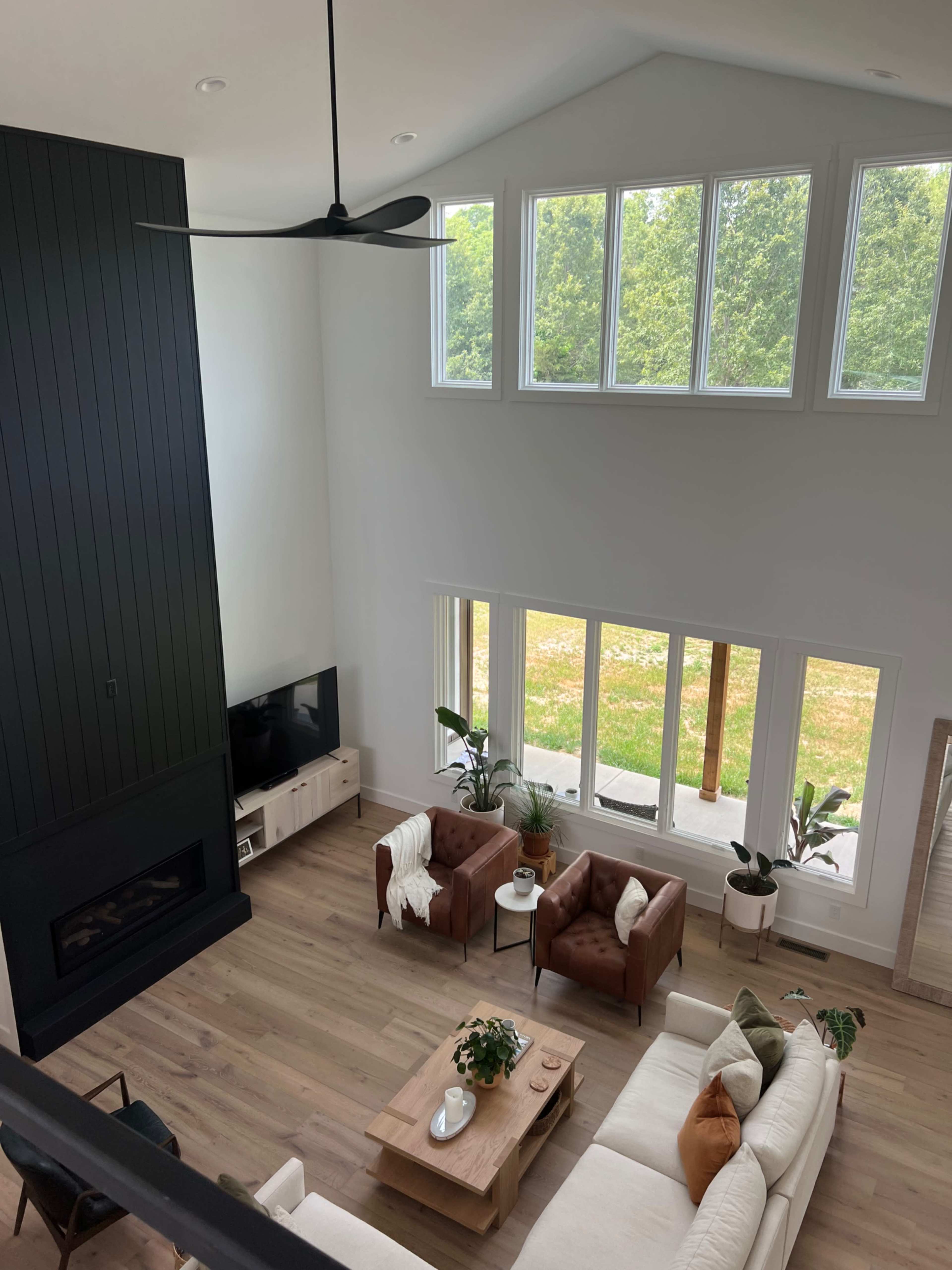 The image shows a modern living room with a large window view overlooking greenery, featuring a fireplace, a coffee table, and seating areas with leather chairs and a sectional sofa.