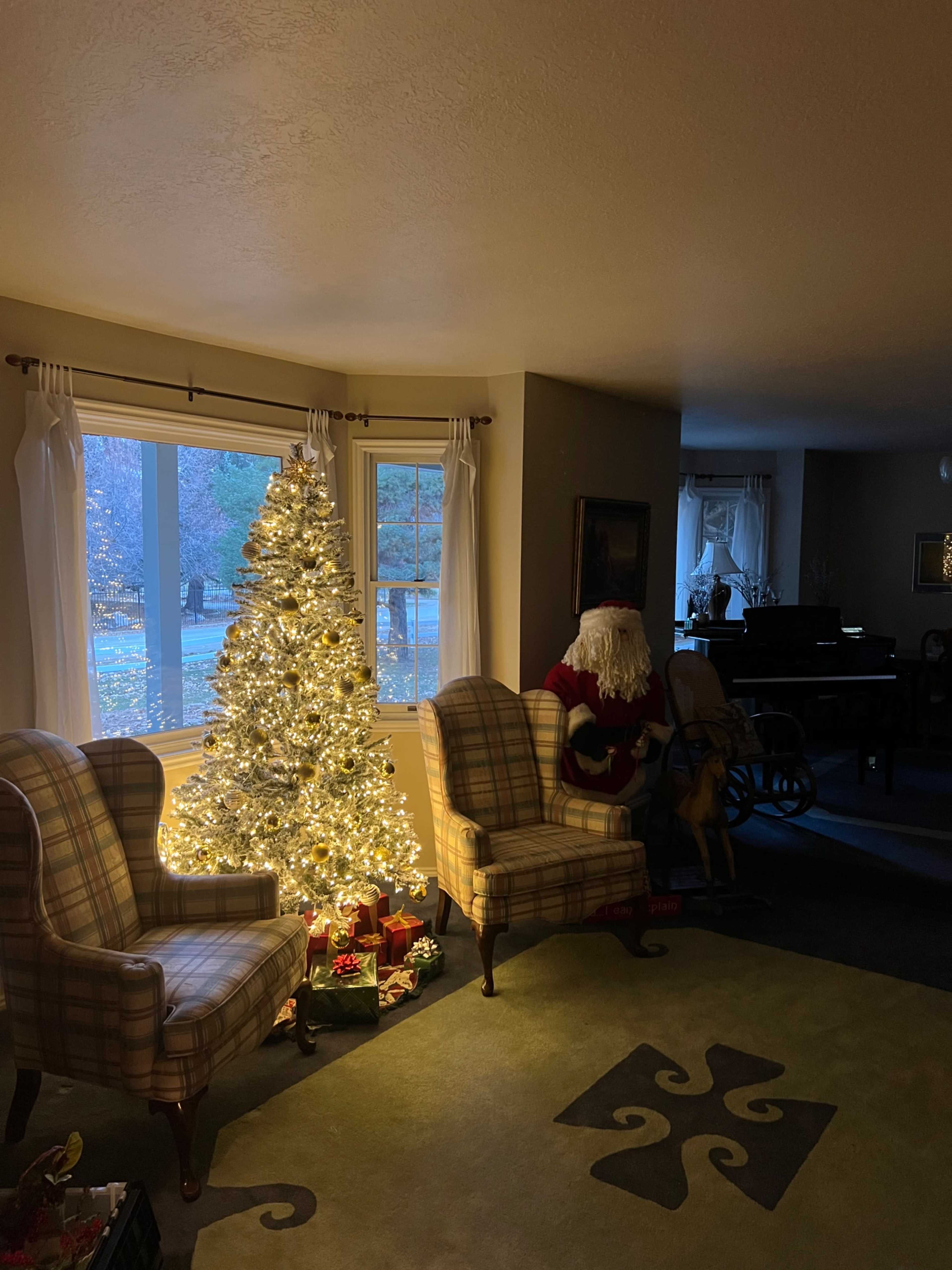 A decorated Christmas tree illuminates a cozy living room with two armchairs and a Santa figure seated nearby.