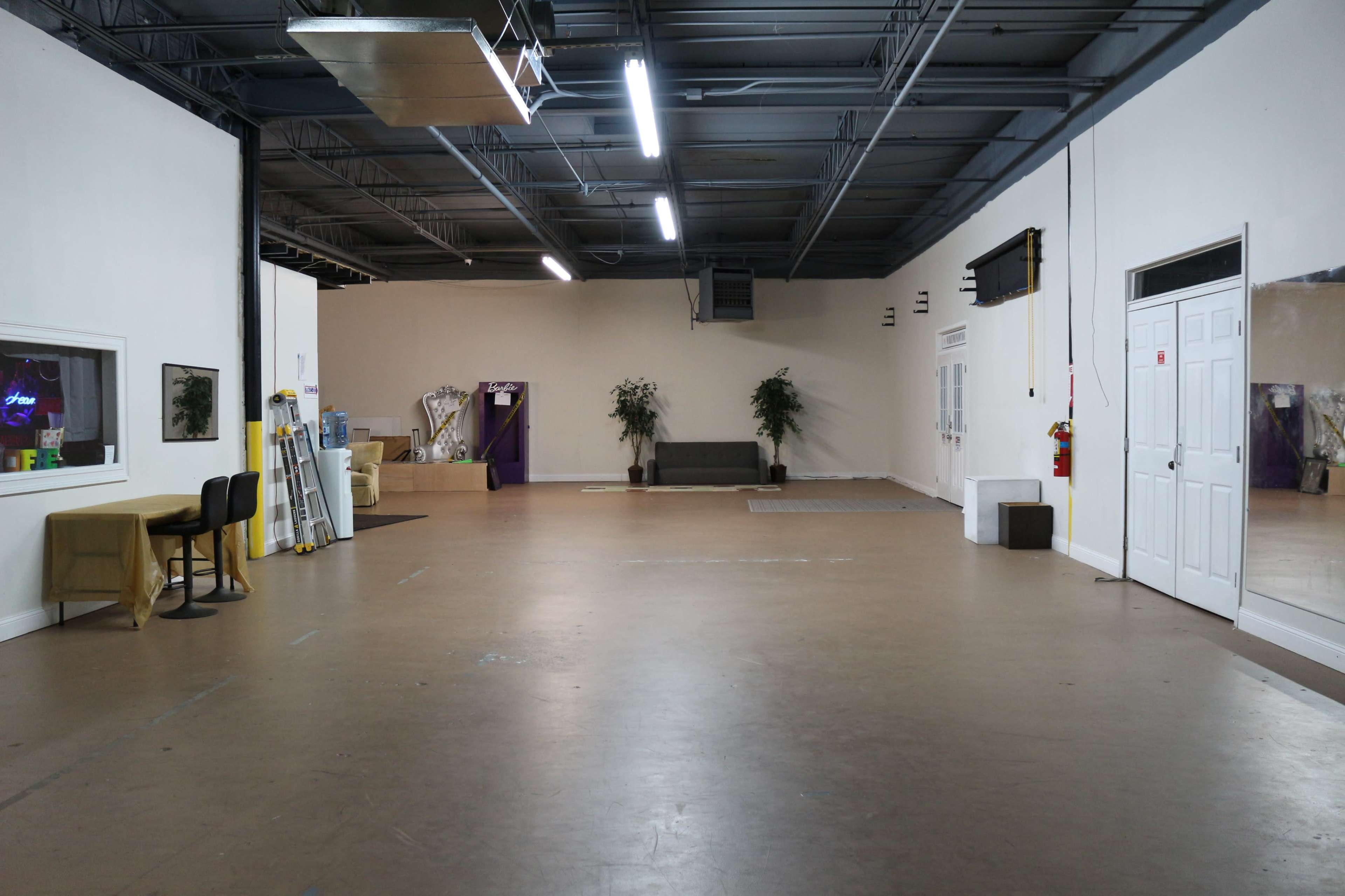 A spacious, empty interior room with a flat floor, neutral-colored walls, and minimal furnishings including a couch and a few plants.