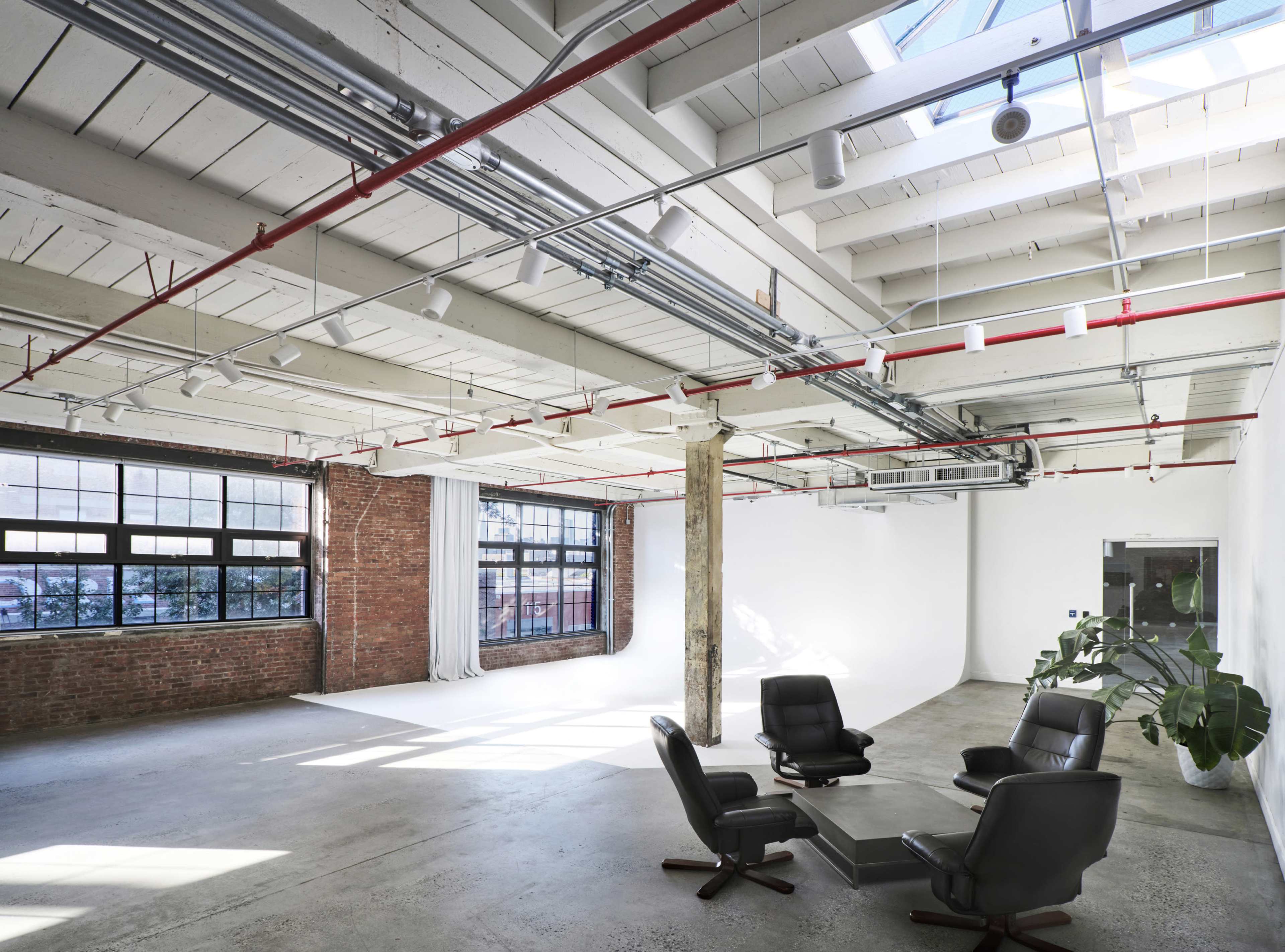 The image shows a spacious, modern interior with exposed brick walls, a concrete floor, and large windows allowing natural light to enter a minimalist meeting area featuring black chairs and a small table.
