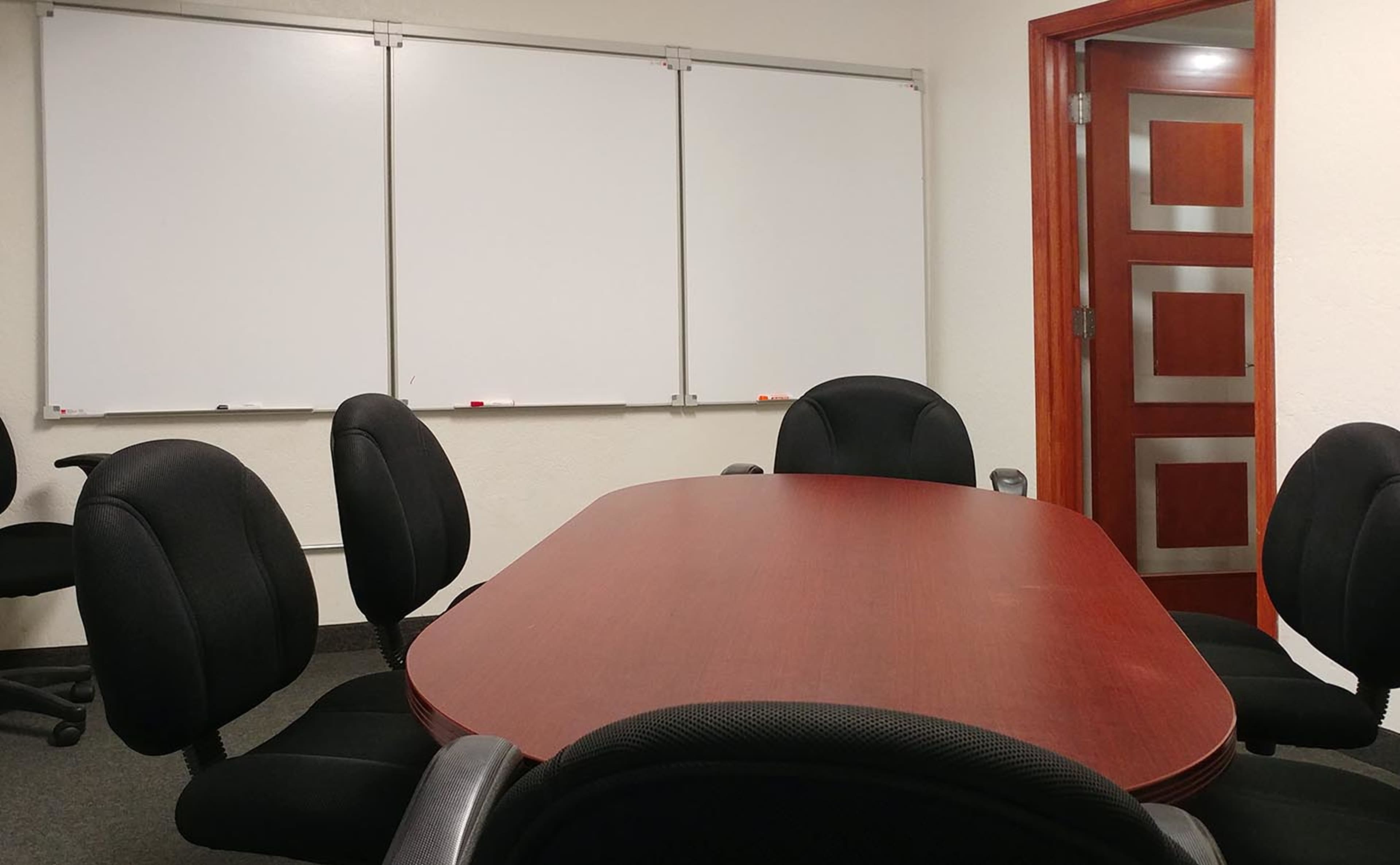 A conference room with a wooden table surrounded by black chairs and empty whiteboards on the wall.