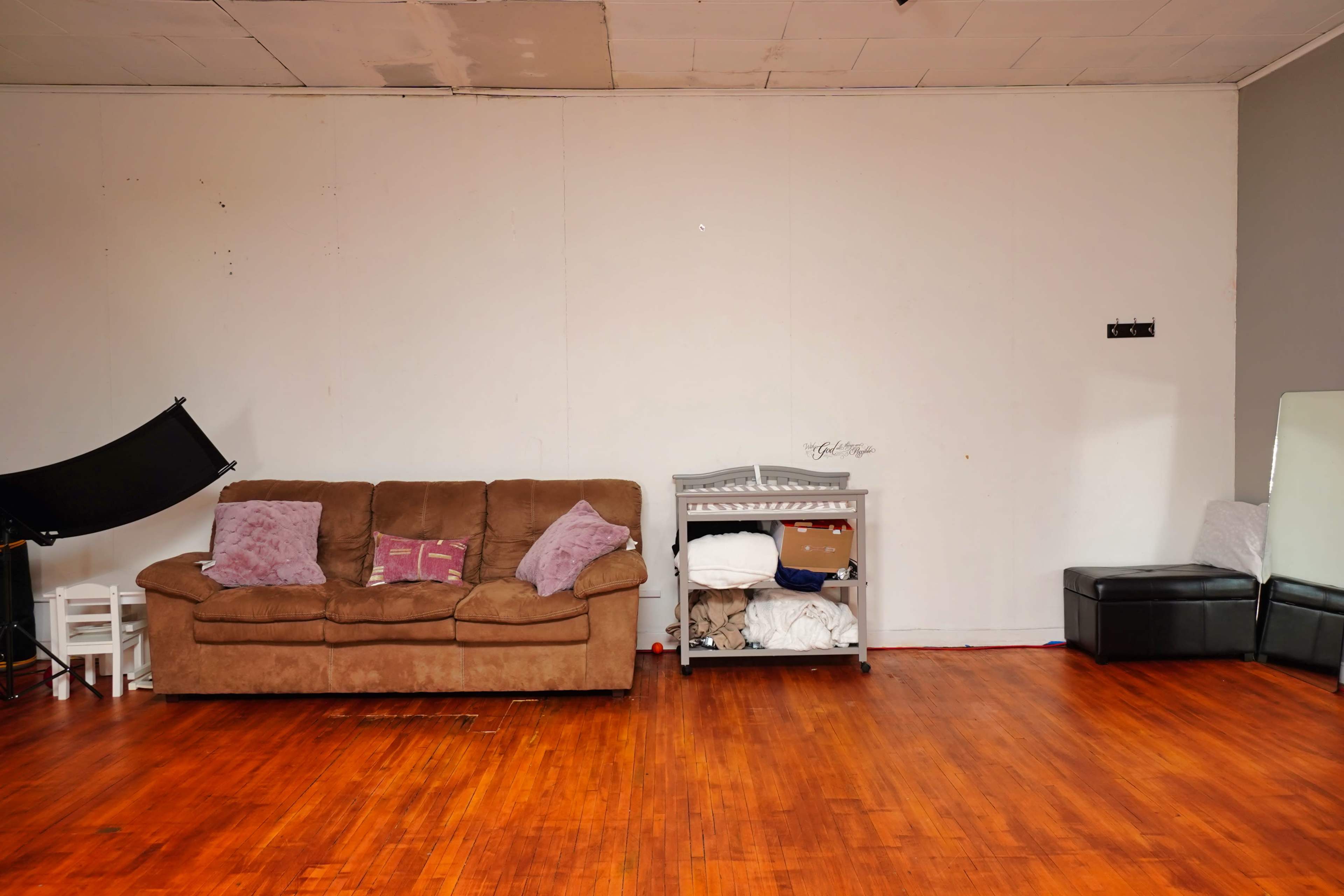 A brown couch is positioned against a white wall, with a storage unit nearby and a black ottoman in the corner of the room.