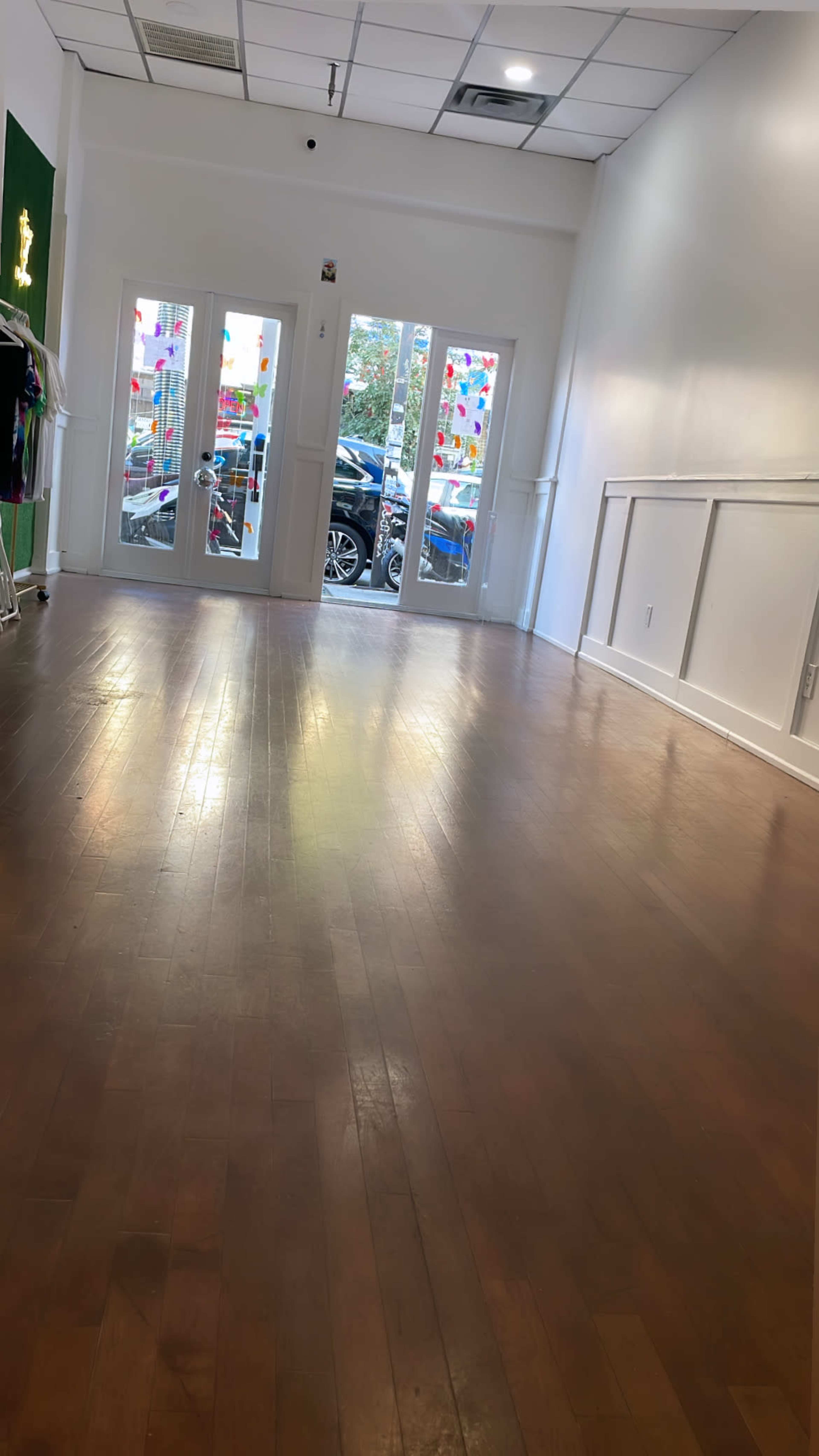 A spacious, empty room with wooden flooring and large glass doors leading to the outdoors, where colorful decorations are visible.