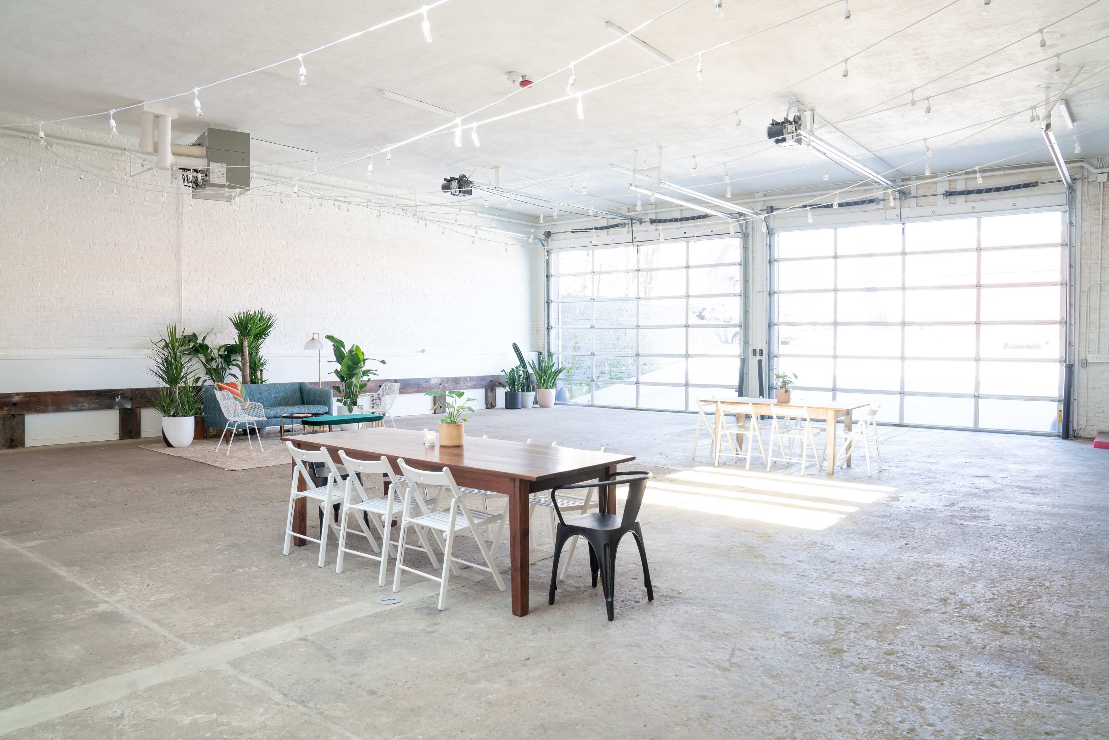 A spacious, well-lit industrial room with large windows, featuring two tables surrounded by chairs and various indoor plants.