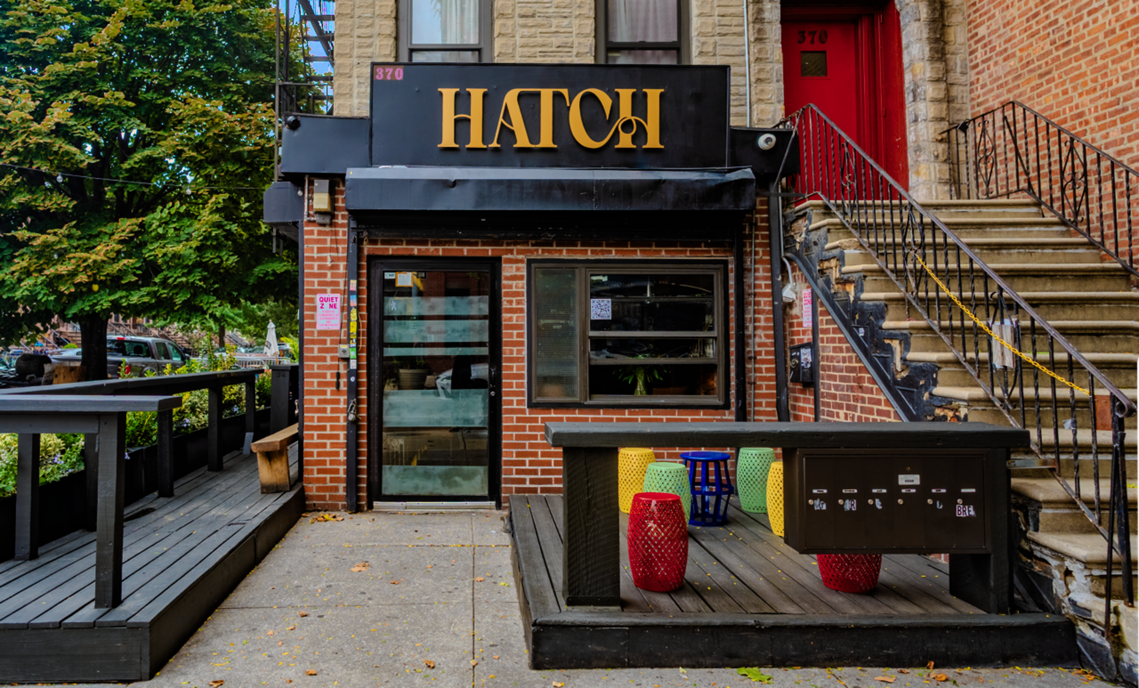 The image shows the entrance of a restaurant called "HATCH," featuring a signage above a glass door and outdoor seating with colorful tables and a mailbox.