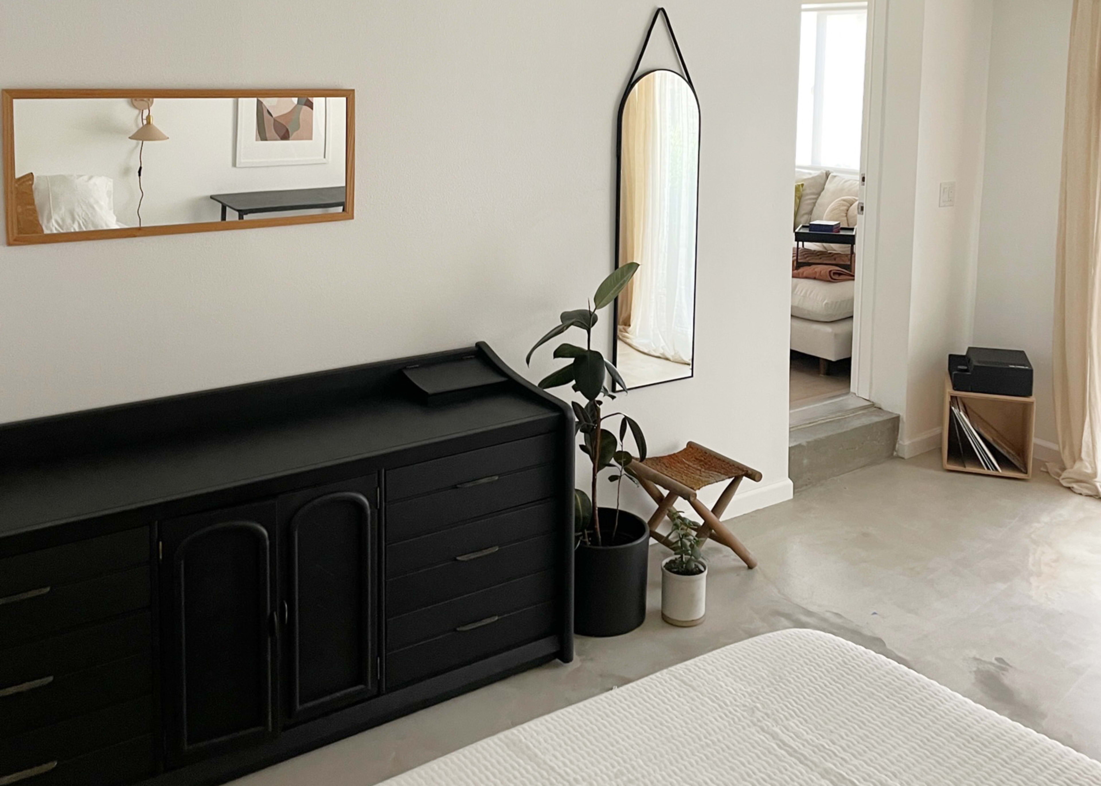 A minimalist bedroom features a black dresser, a mirror on the wall, a potted plant, and a glimpse of a living area through an open doorway.
