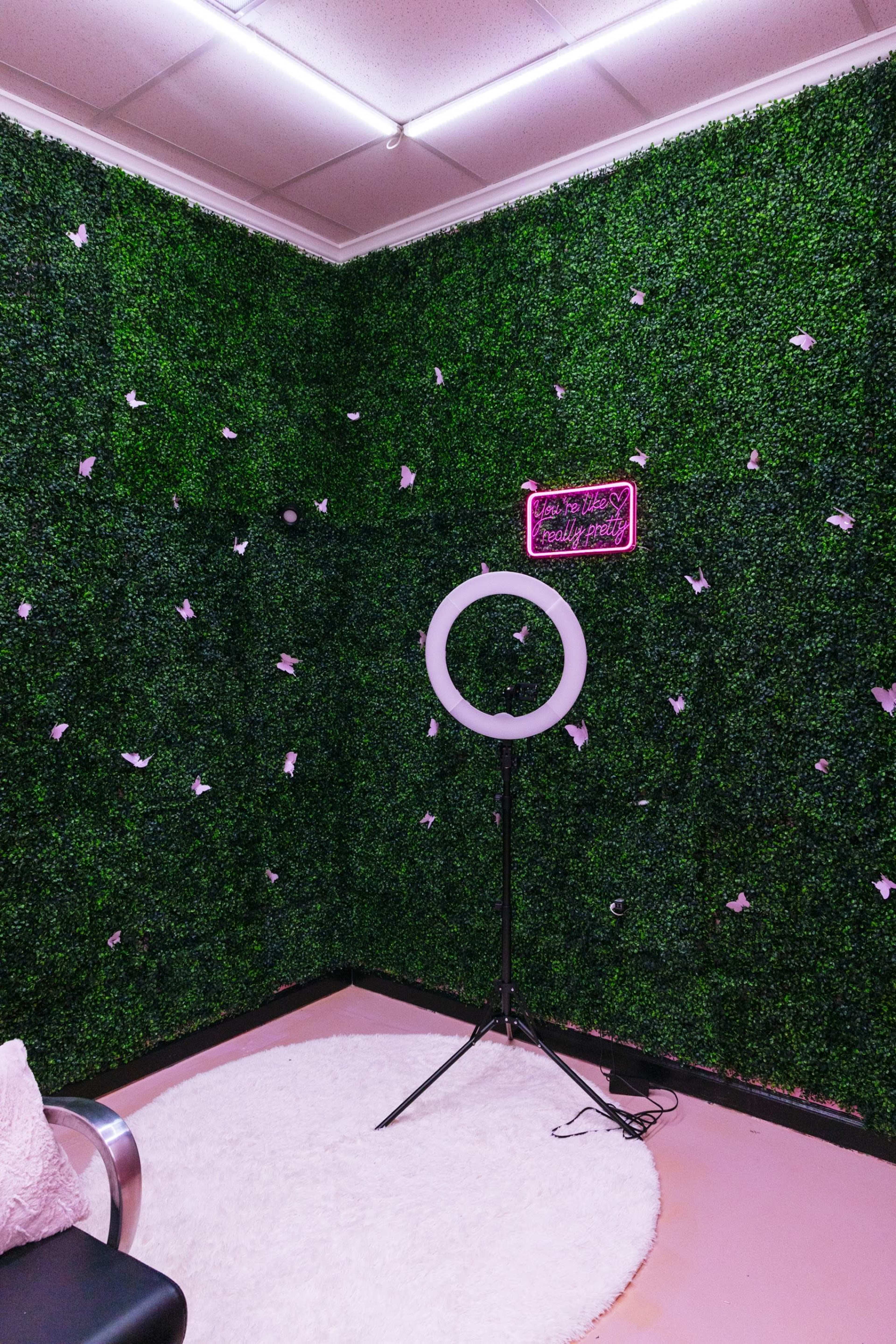 The image shows a corner of a room with a green artificial hedge wall, a circular light stand, a round rug on the floor, and a neon sign that reads "You're beautiful, don't forget."