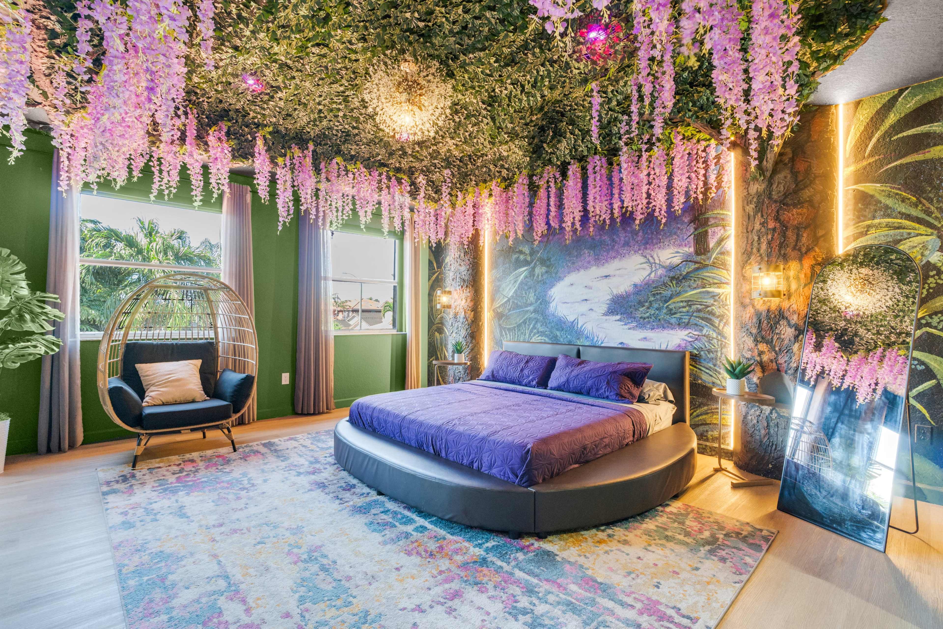 A large bedroom features a circular bed with purple bedding, surrounded by a ceiling adorned with hanging greenery and purple flowers, complemented by a vibrant mural and a cozy seating area.