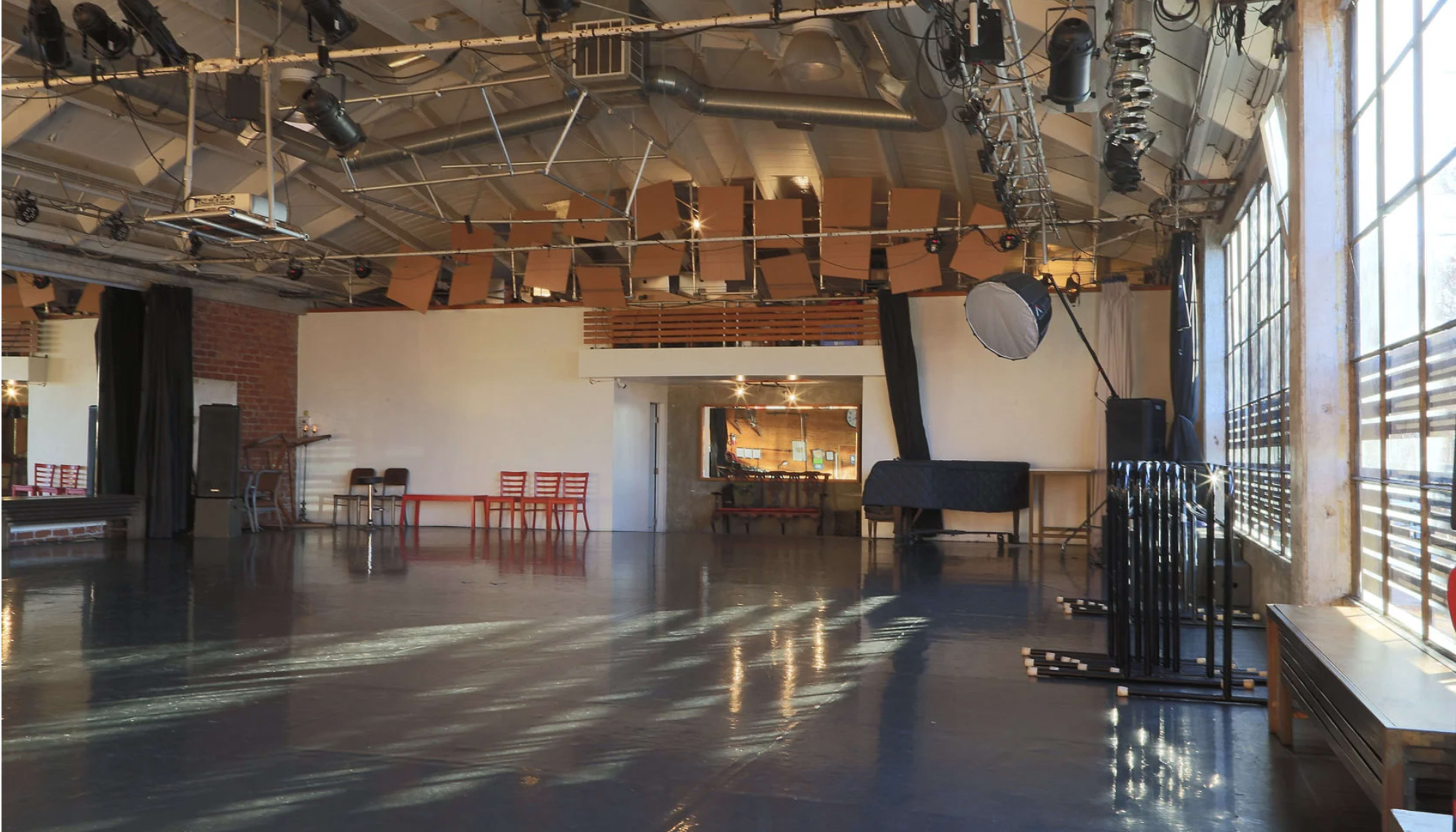 The image shows a spacious, well-lit studio with a polished floor, large windows, and several red chairs arranged near a mirror.