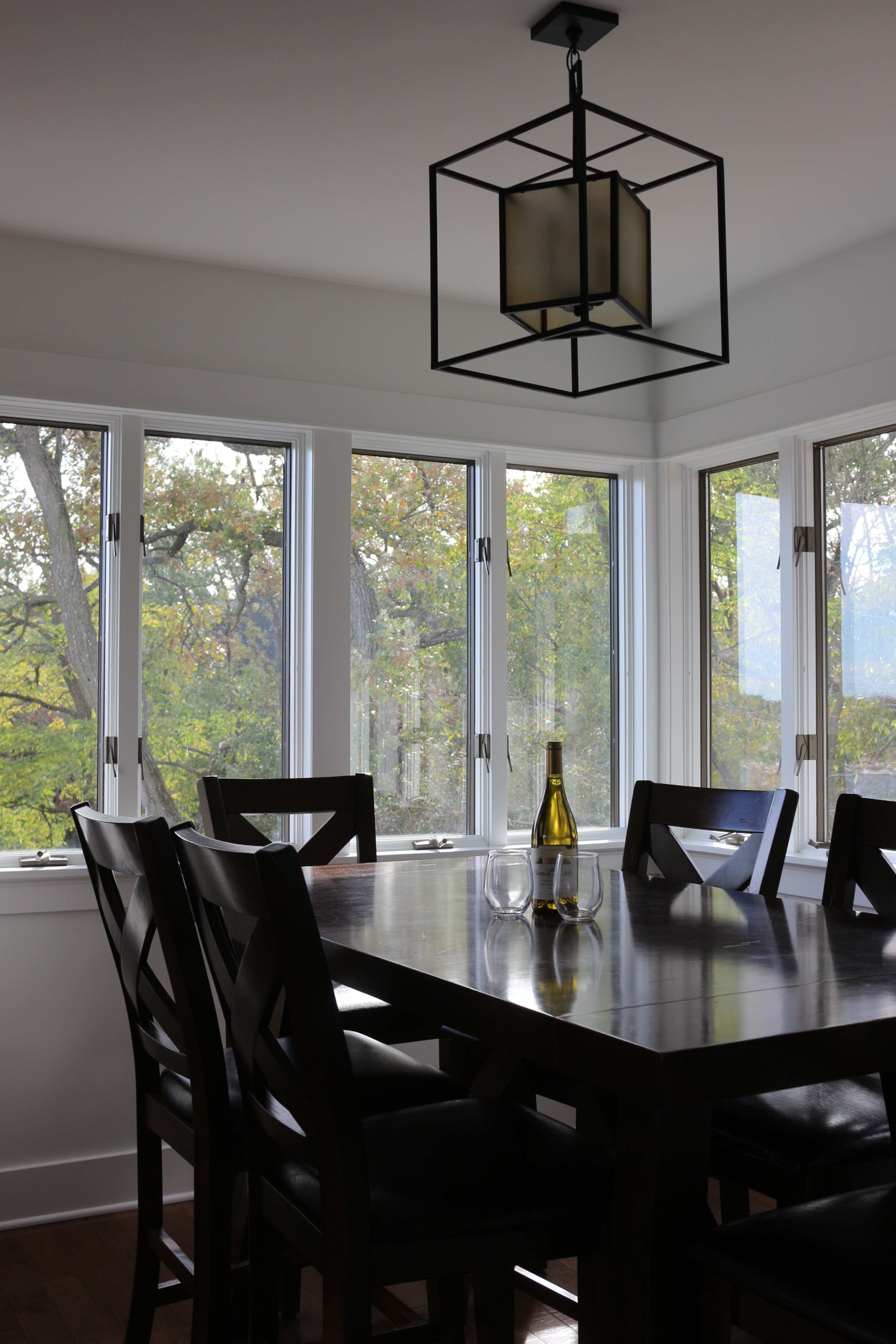 A modern dining area features a wooden table set for two, with a bottle of wine and glasses, illuminated by a geometric light fixture and surrounded by large windows showcasing greenery outside.